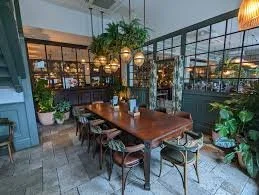 Interior of a cozy restaurant with a wooden table, surrounded by chairs, large windows, green plants, and ambient lighting.
