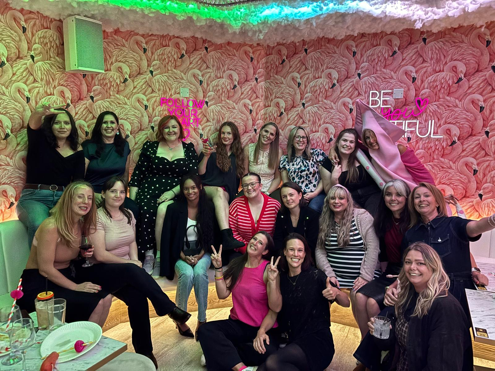 A group of women gathered in a decorated room with flamingo wall wallpaper, neon signs, and a pink flamingo costume. They are smiling, sitting, and standing, celebrating together with drinks and party accessories.