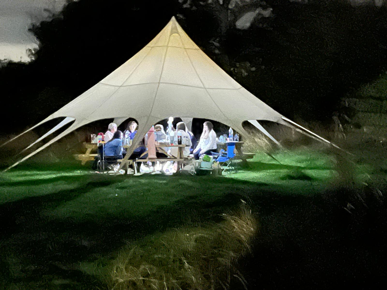A nighttime outdoor gathering under a large beige tent with people sitting at tables, eating, drinking, and socializing on a grassy area.