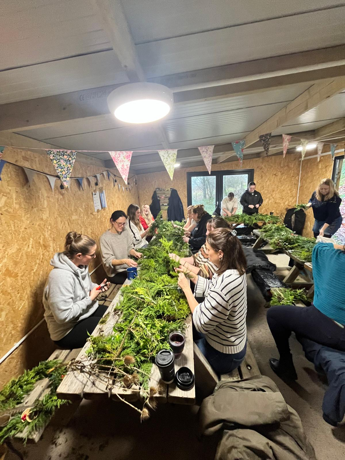 People sitting at a long table inside a wooden room, engaging in a craft activity with greenery. Decorations include colorful triangular bunting hanging from the ceiling.