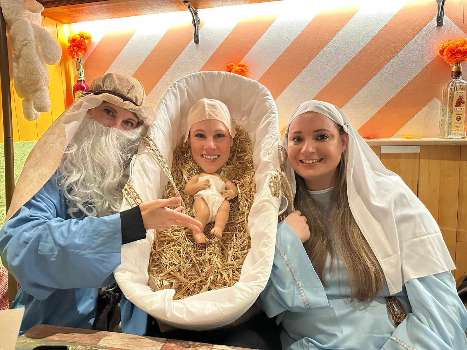 Three women dressed as biblical figures with one woman holding a baby doll, all smiling, in a cozy, decorated room.