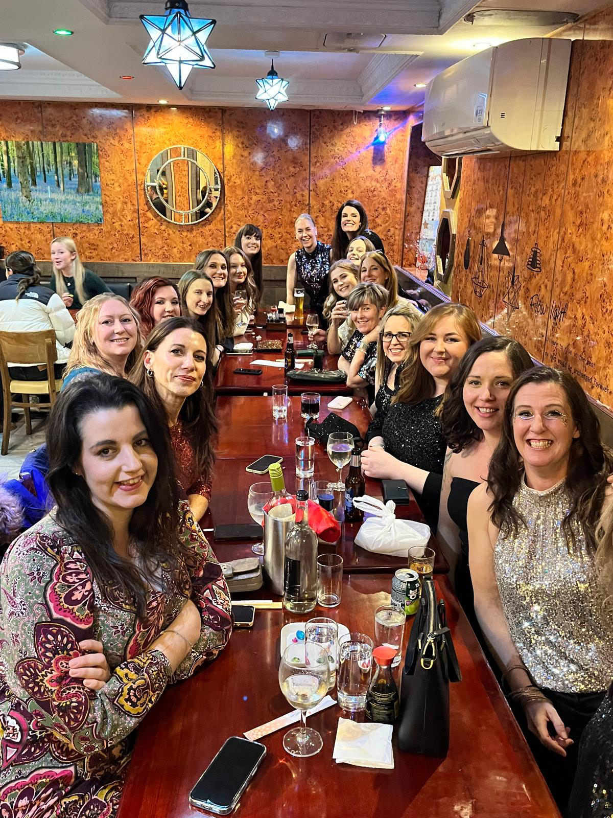 Group of women gathered around a long wooden table at a restaurant, smiling and enjoying drinks, with colorful lighting and decorative wall hangings.