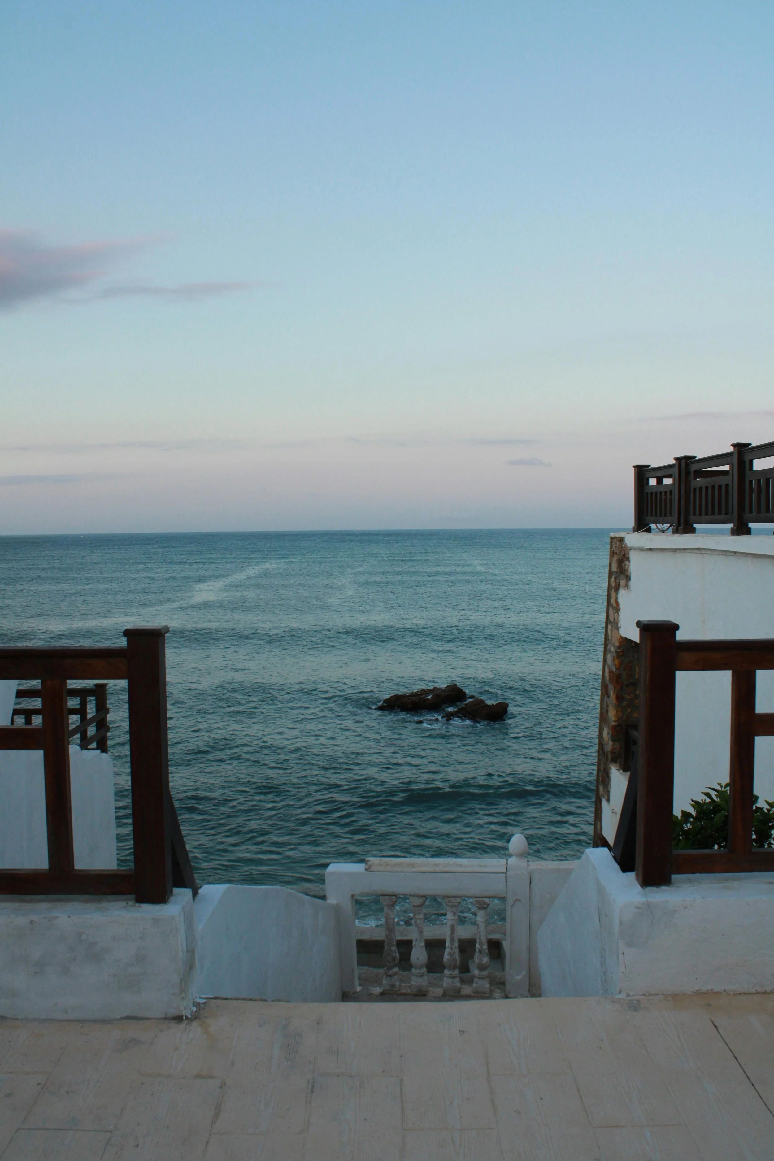 View of the ocean from a balcony or terrace with white walls, wooden railings, and a small set of stairs leading down to the water, under a partly cloudy sky.