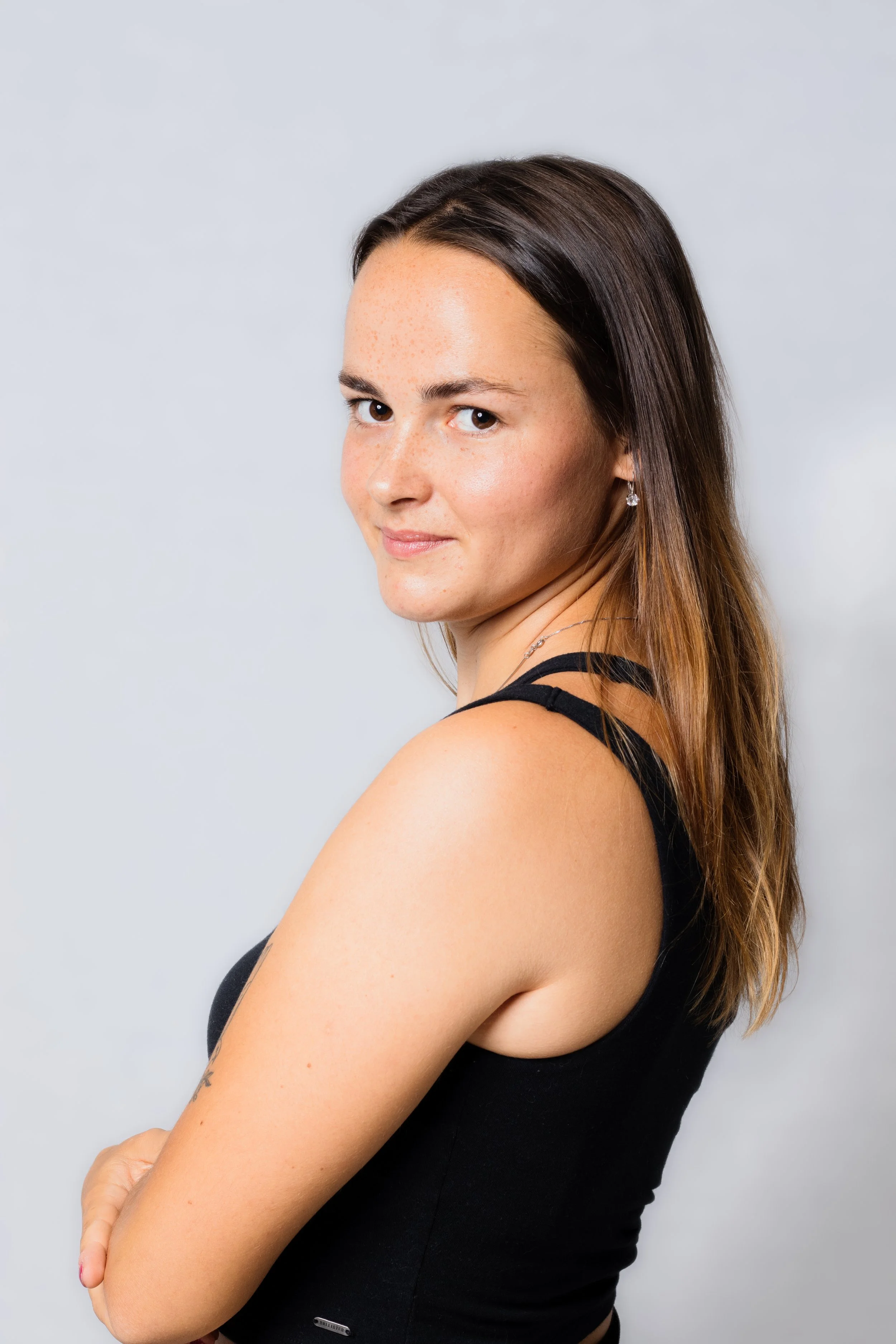 A woman with long brown hair, wearing a black sleeveless top, standing against a light gray background.