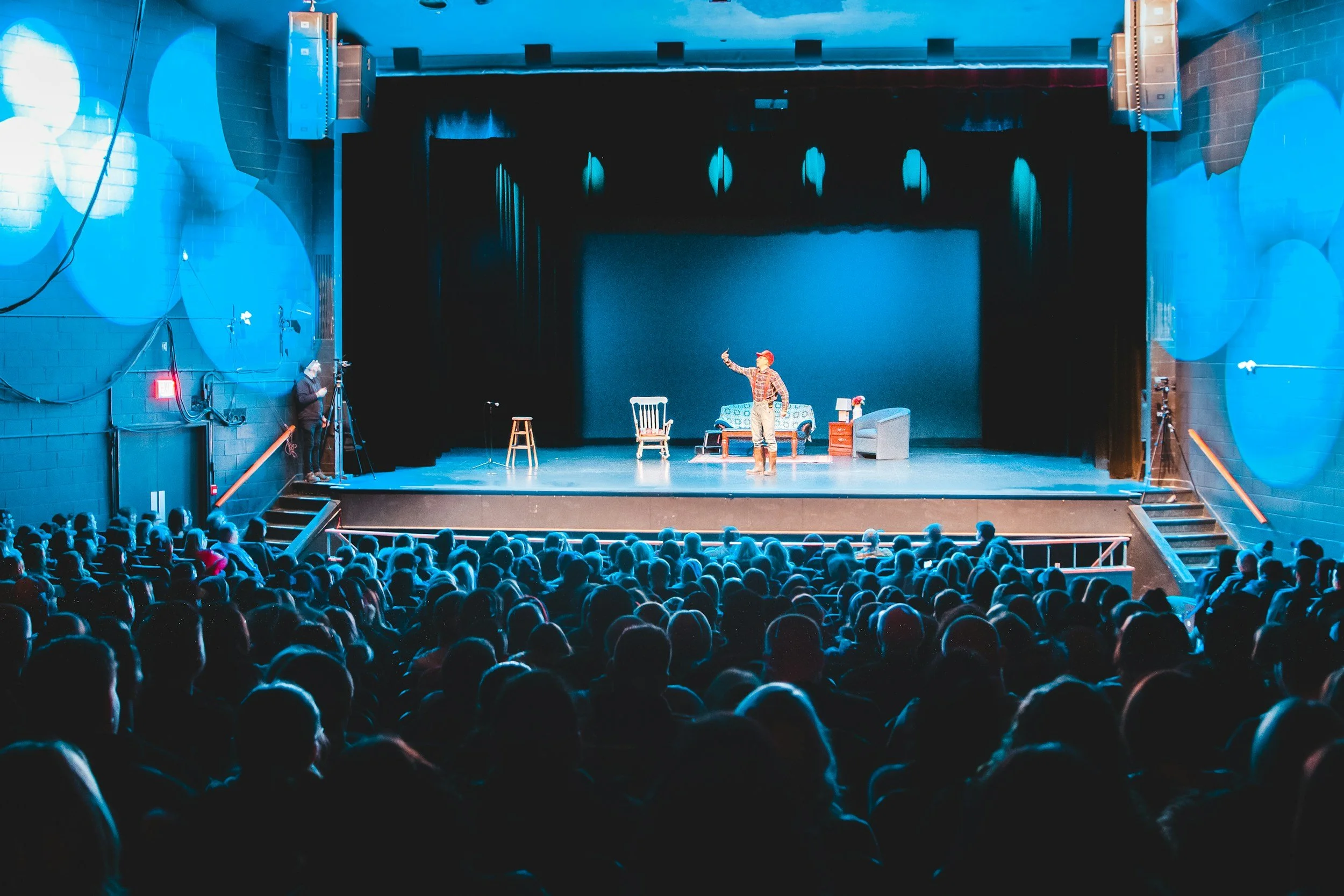 A theater stage with a performer in front of an audience. The stage has furniture including chairs, a table with a lamp, and a sofa. The performer is wearing a red cap, a plaid shirt, and tan pants, and is gesturing with their right hand.