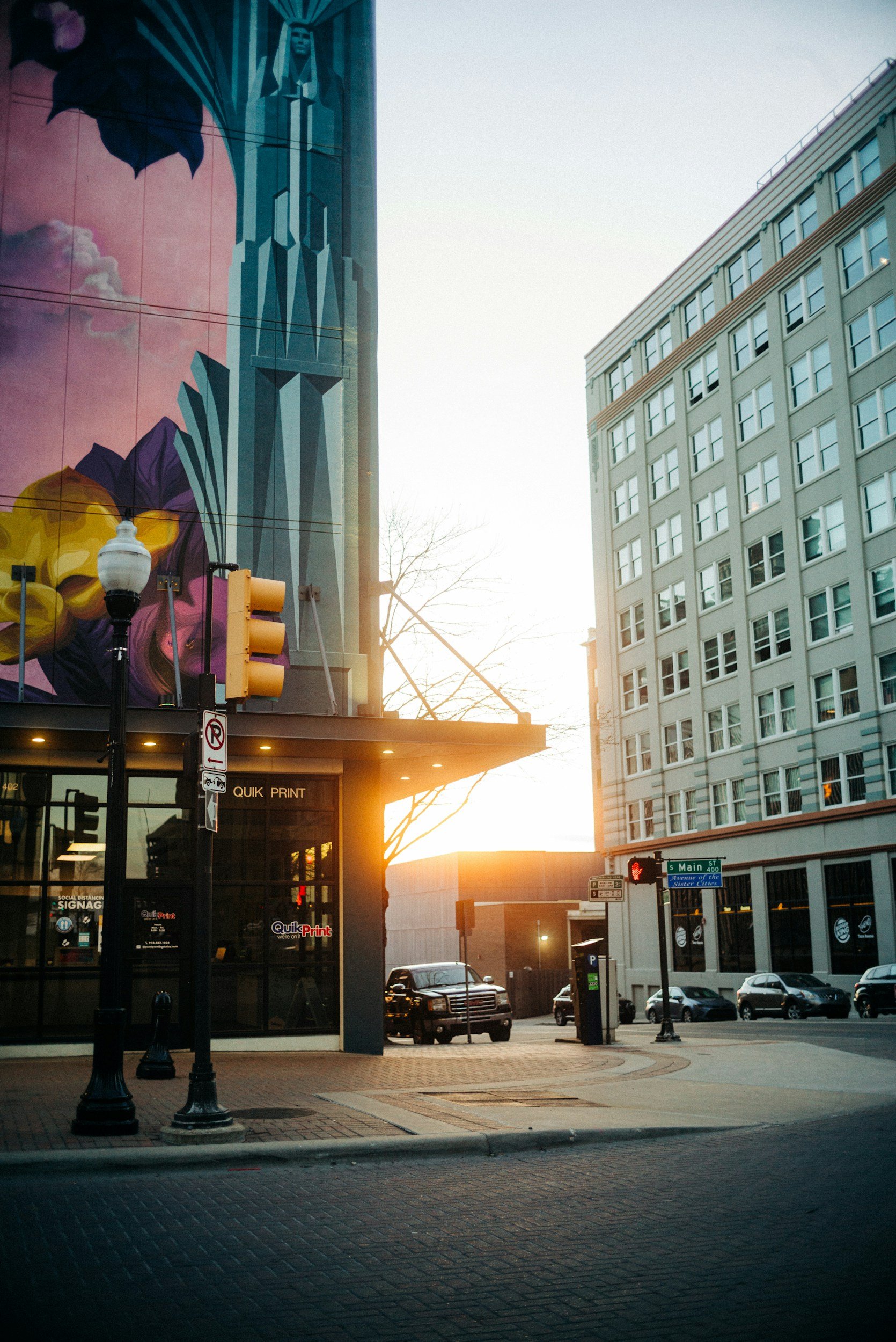 City street scene at sunset with a colorful mural on a building, parked cars, traffic lights, and a street sign indicating Main Street.
