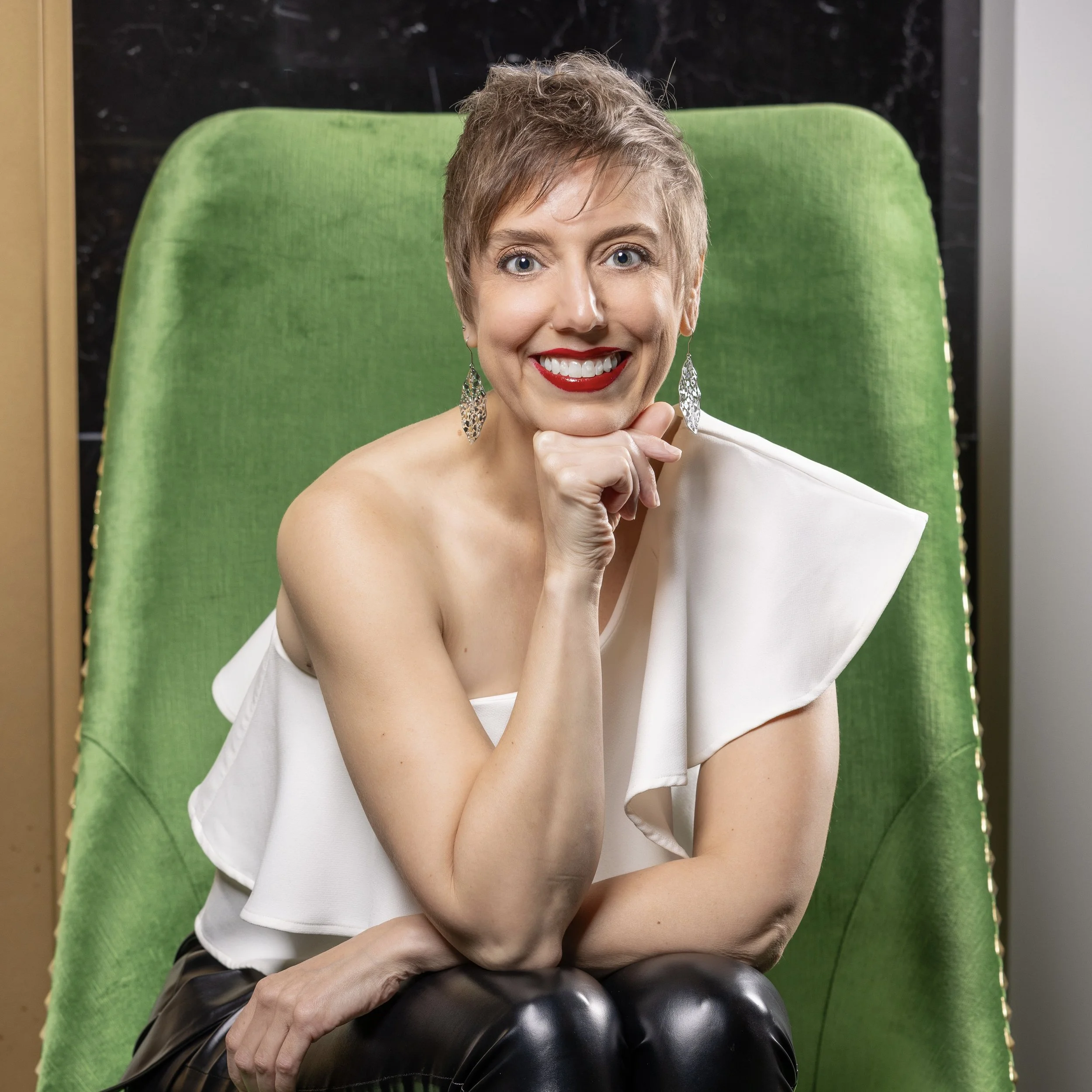A woman with short, light brown hair and bright red lipstick smiling, sitting in a green chair, resting her chin on her hand, wearing a white off-shoulder top and large dangling earrings.