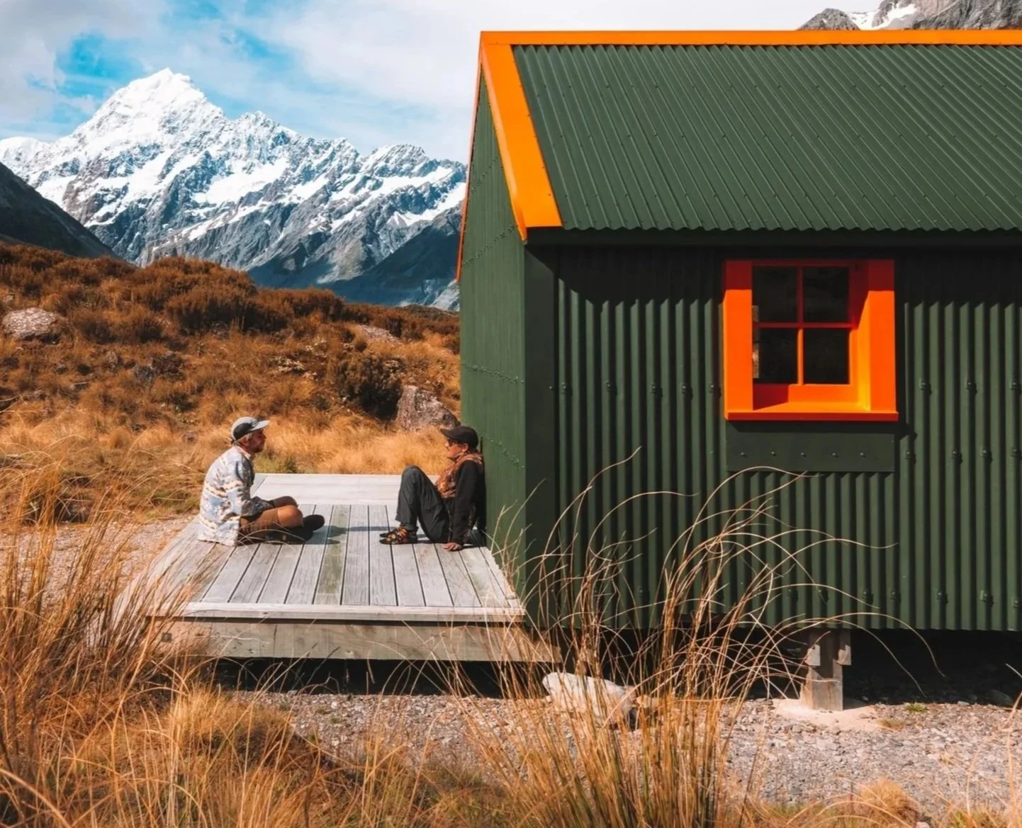 Two people sitting cross-legged on a wooden deck, facing each other, in front of a green building with orange window framing in a mountain landscape with snow-capped peaks and dry brownish grass.