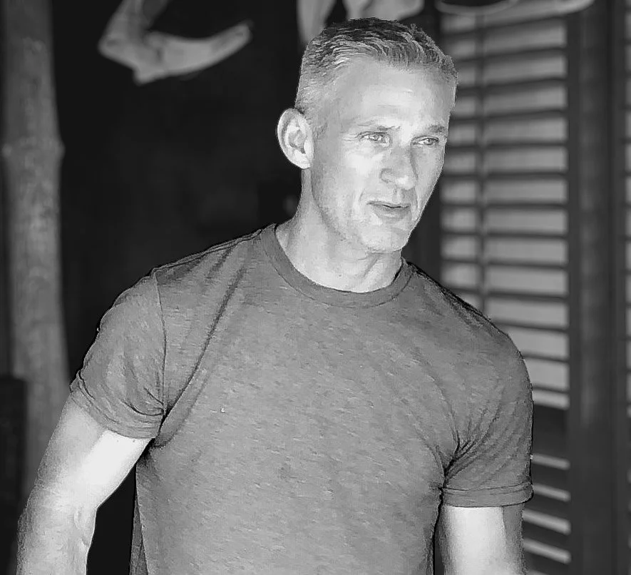 A black and white photo of a man with short hair, wearing a fitted t-shirt, standing indoors in front of a wooden slatted wall.