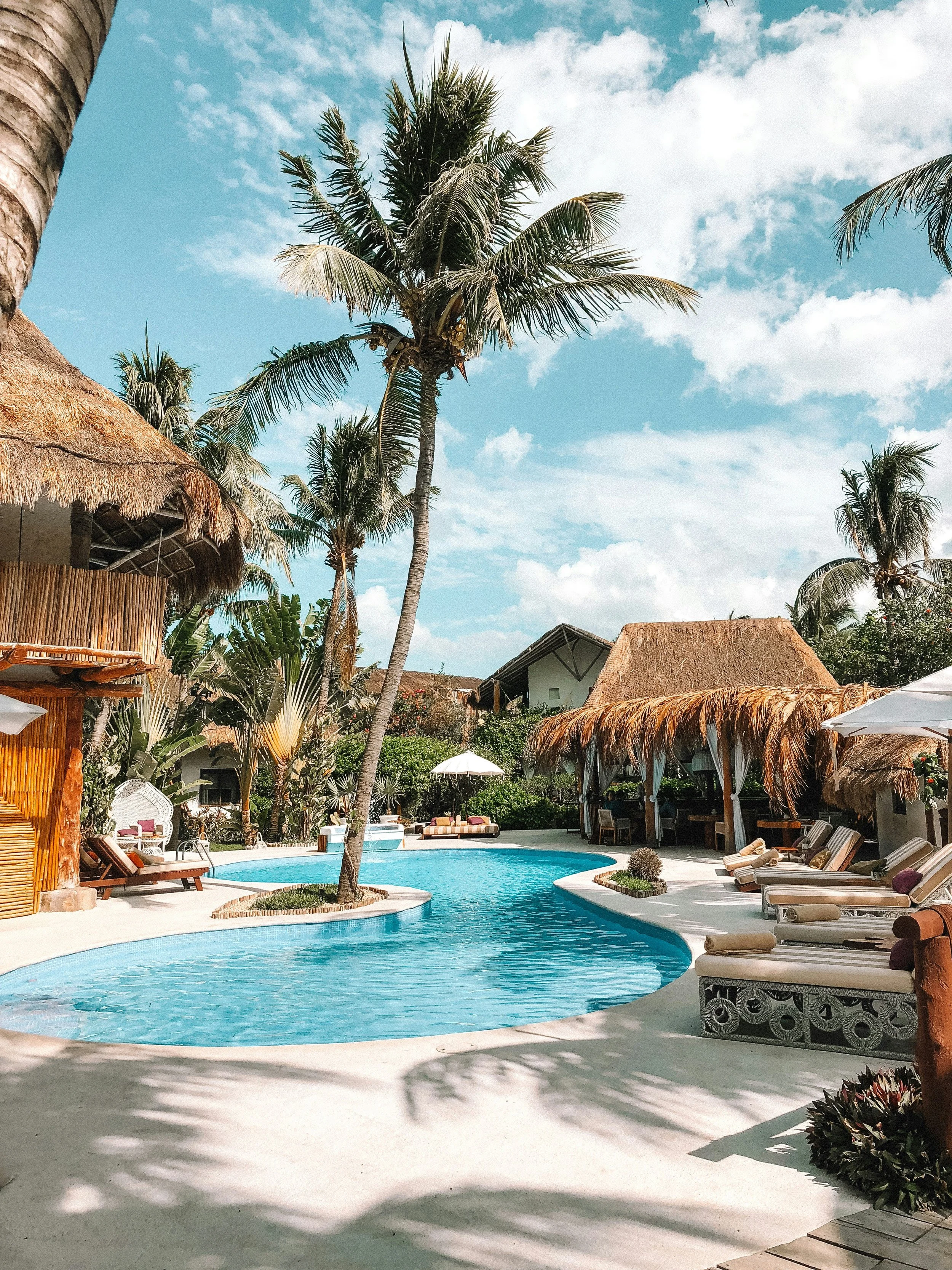 A tropical resort pool area with palm trees, thatched-roof huts, poolside lounges, and a bright blue sky with scattered clouds.