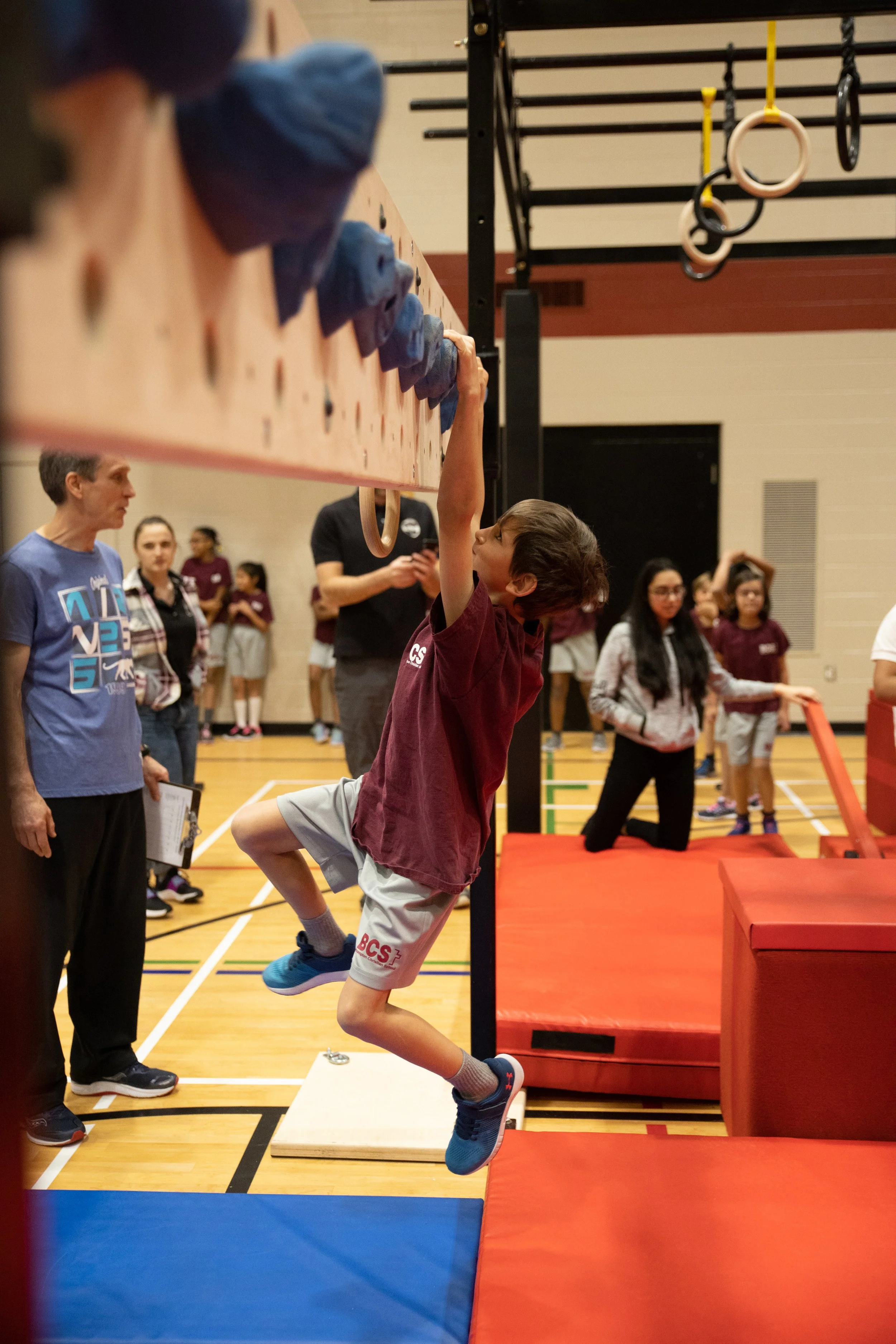 A boy in a maroon T-shirt and gray shorts is hanging from a climbing wall, holding onto a ring with one hand. Several people are observing in a gymnasium, with some waiting in line and others watching the activity.