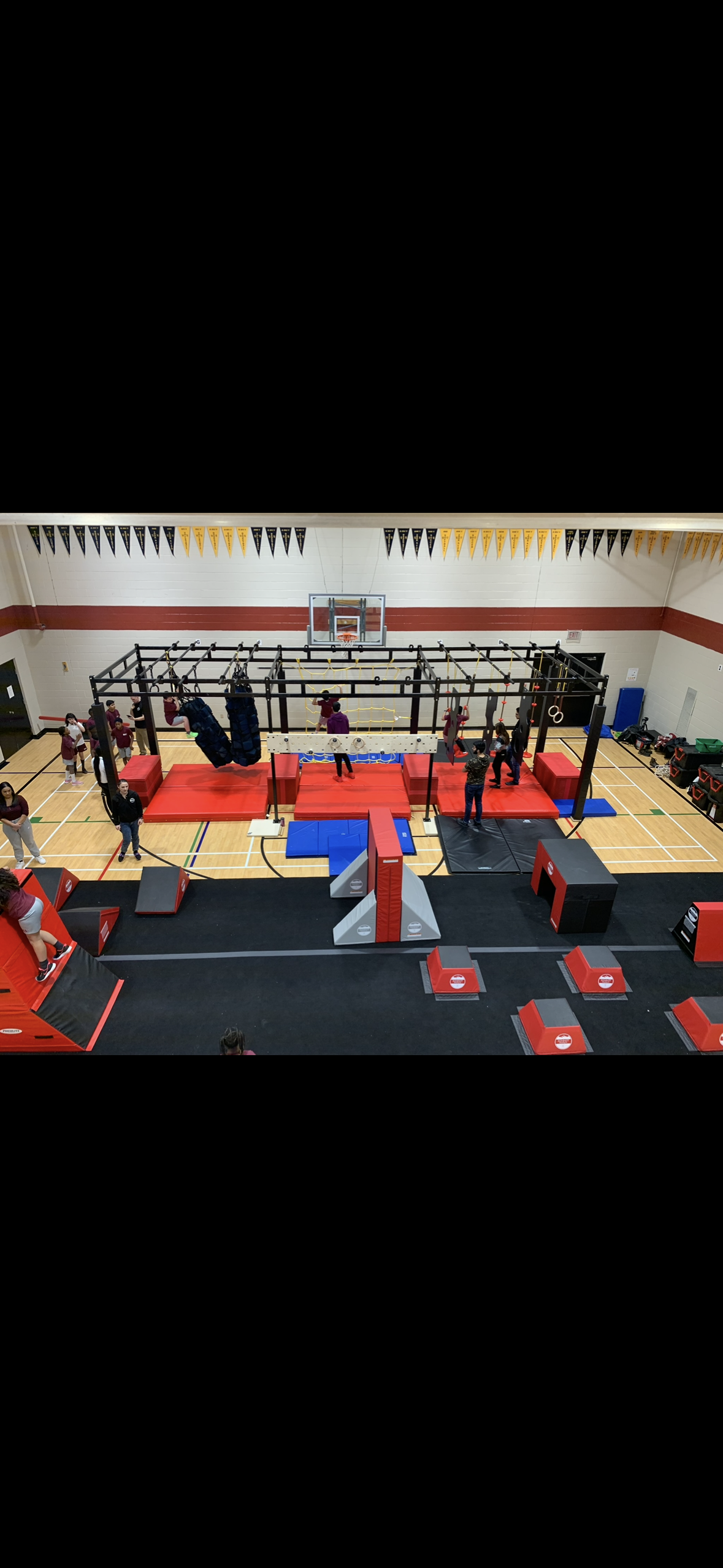 Indoor gym with children and adults playing on obstacle course and climbing structures, basketball hoop, gym mats, and gymnastic rings.