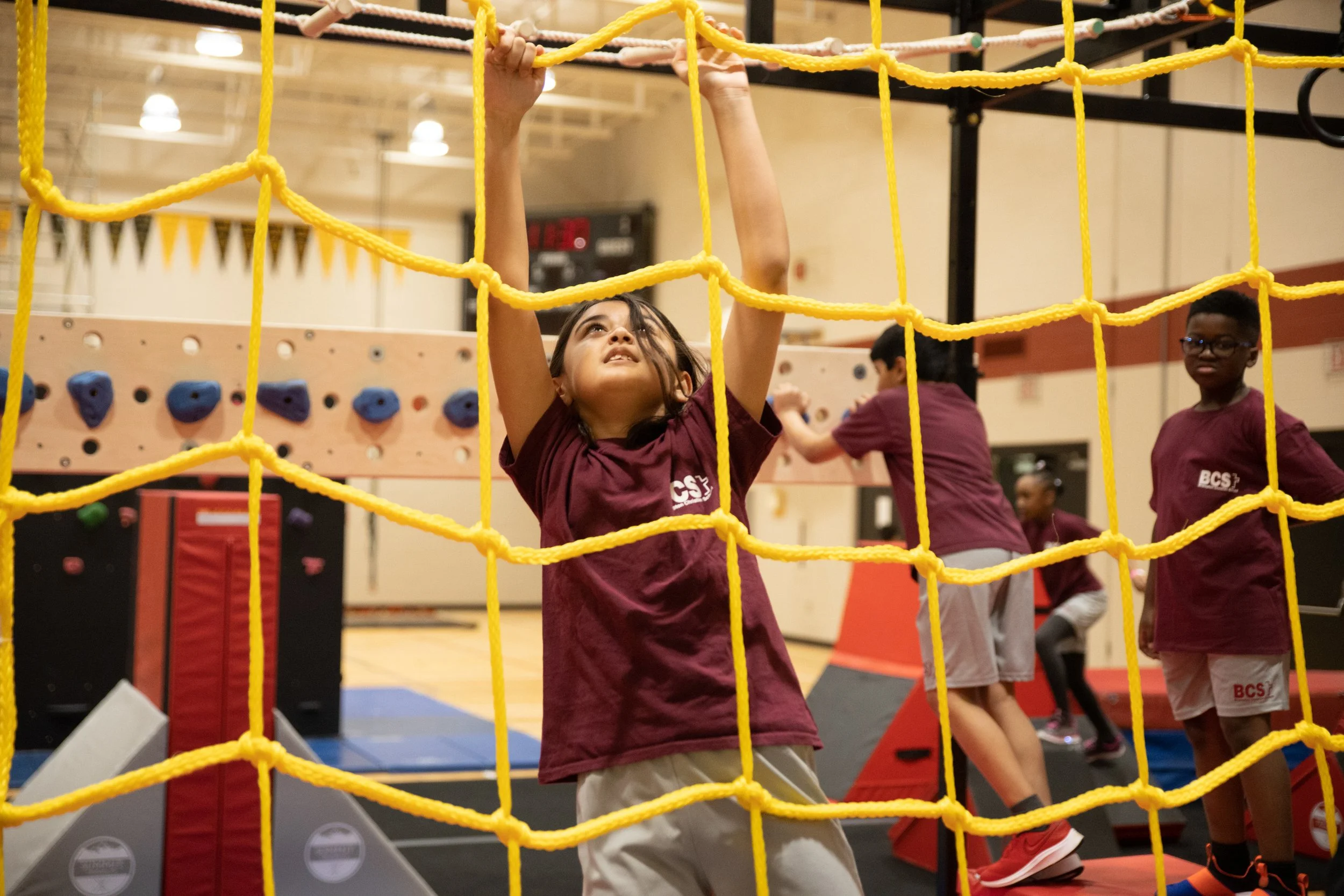 A young girl reaching up and grabbing a yellow climbing rope in an indoor gym, with other children in the background, some climbing and others standing.