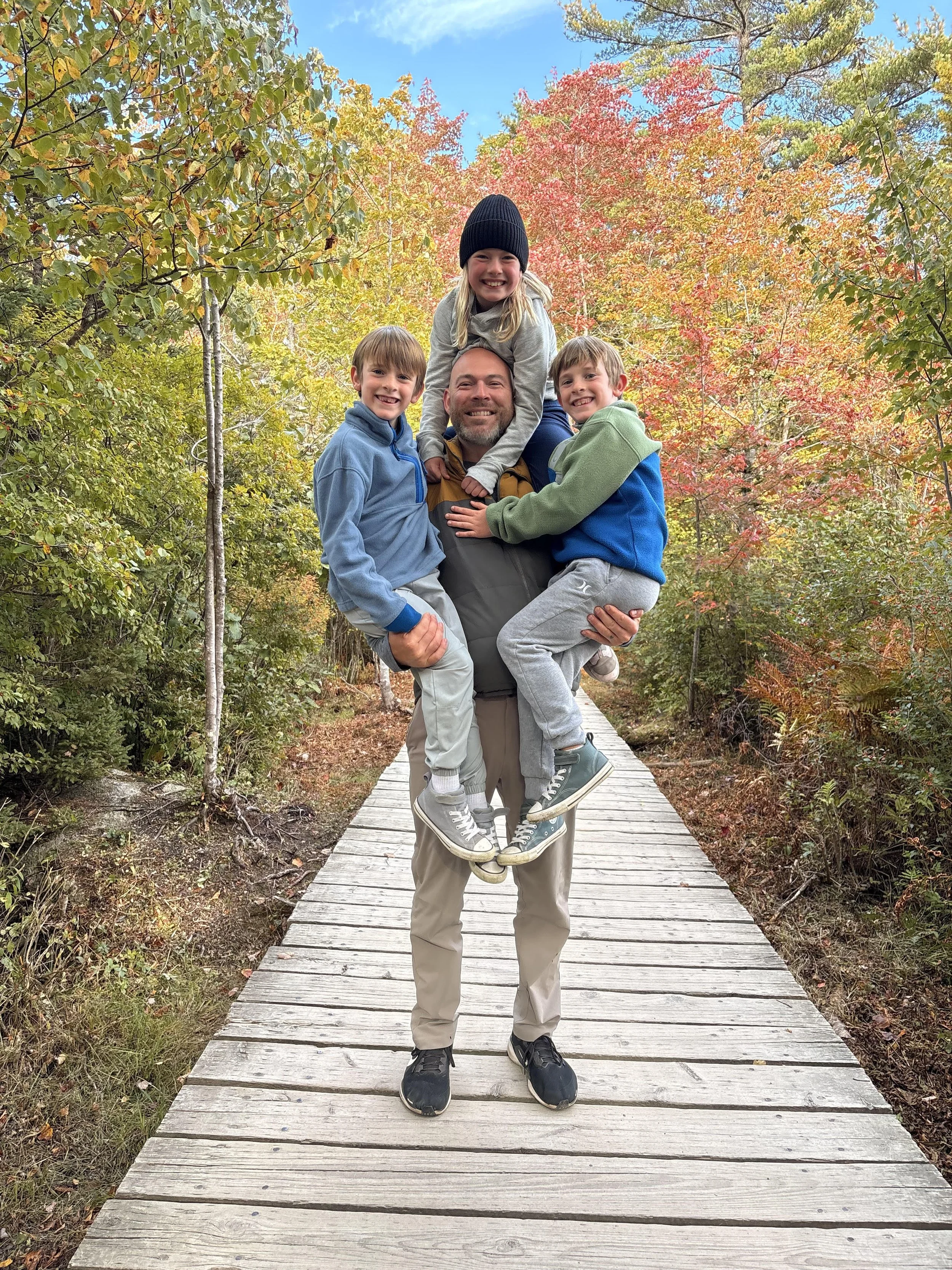 A man holding two boys and one girl on his shoulders on a wooden trail surrounded by colorful fall foliage.