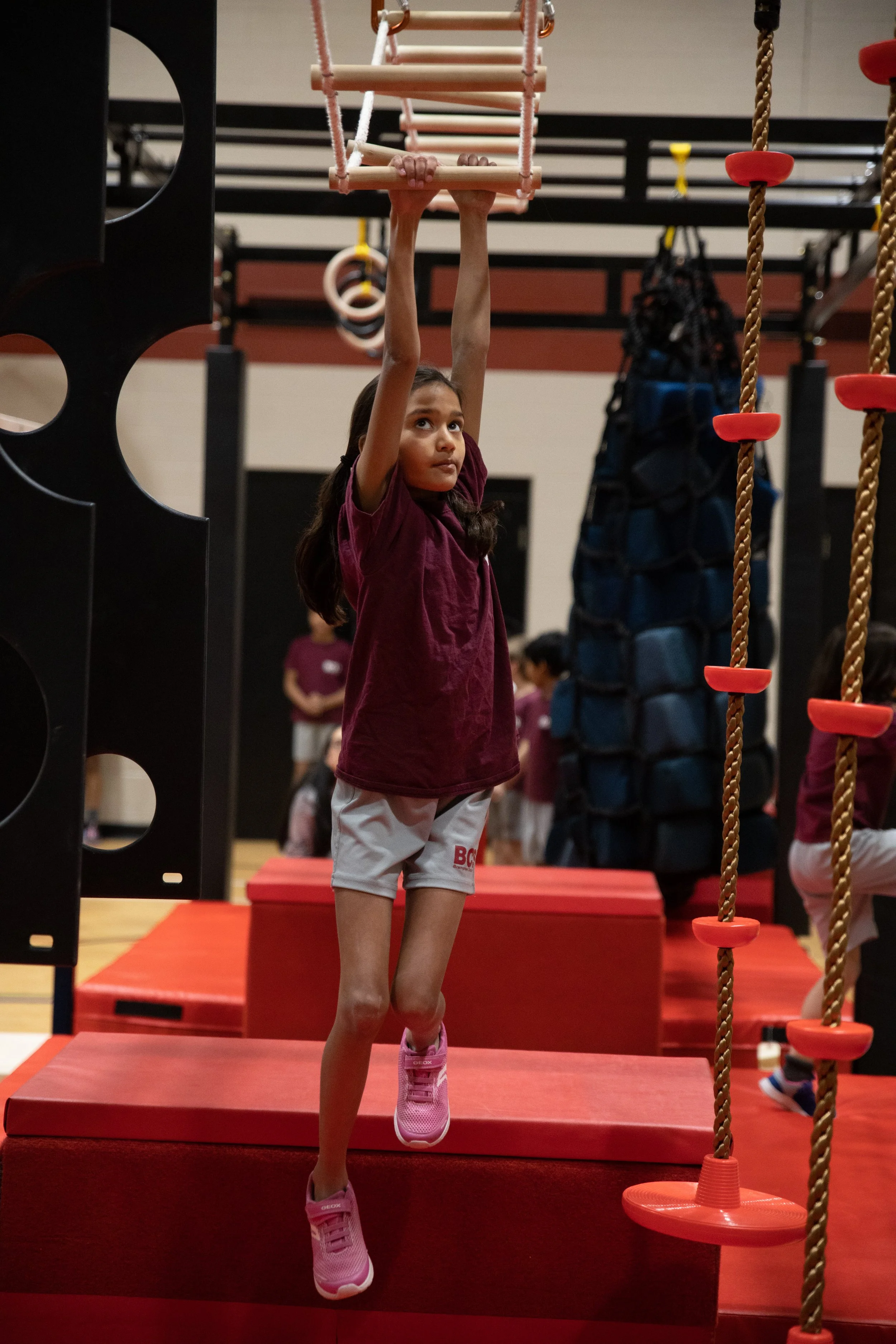 A young girl in a maroon shirt and shorts is hanging from a pink climbing frame in an indoor gym or playground, with other children in the background.