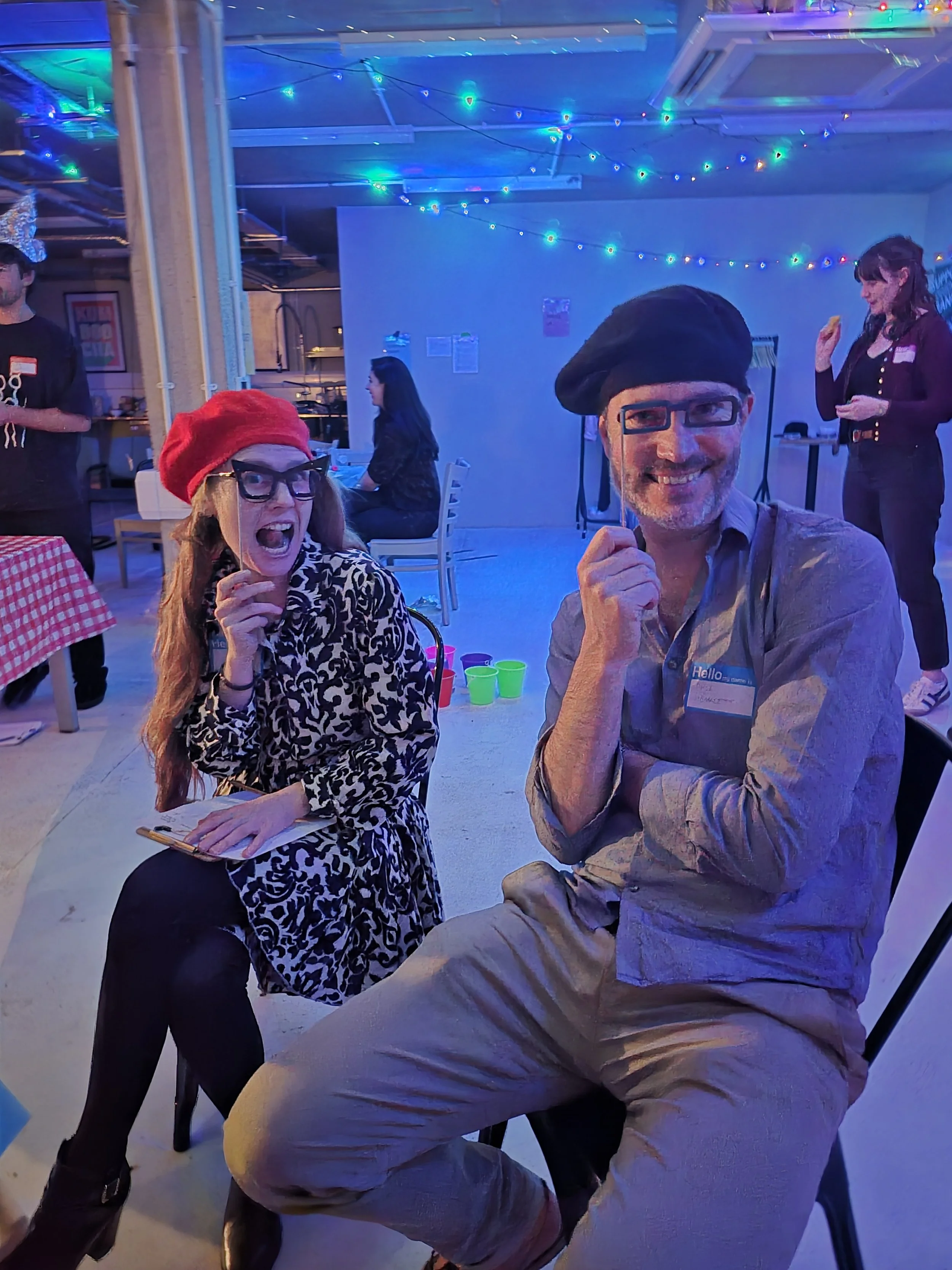 Two people sitting and smiling at a party with festive lights, wearing berets and glasses, surrounded by other guests and decorations.