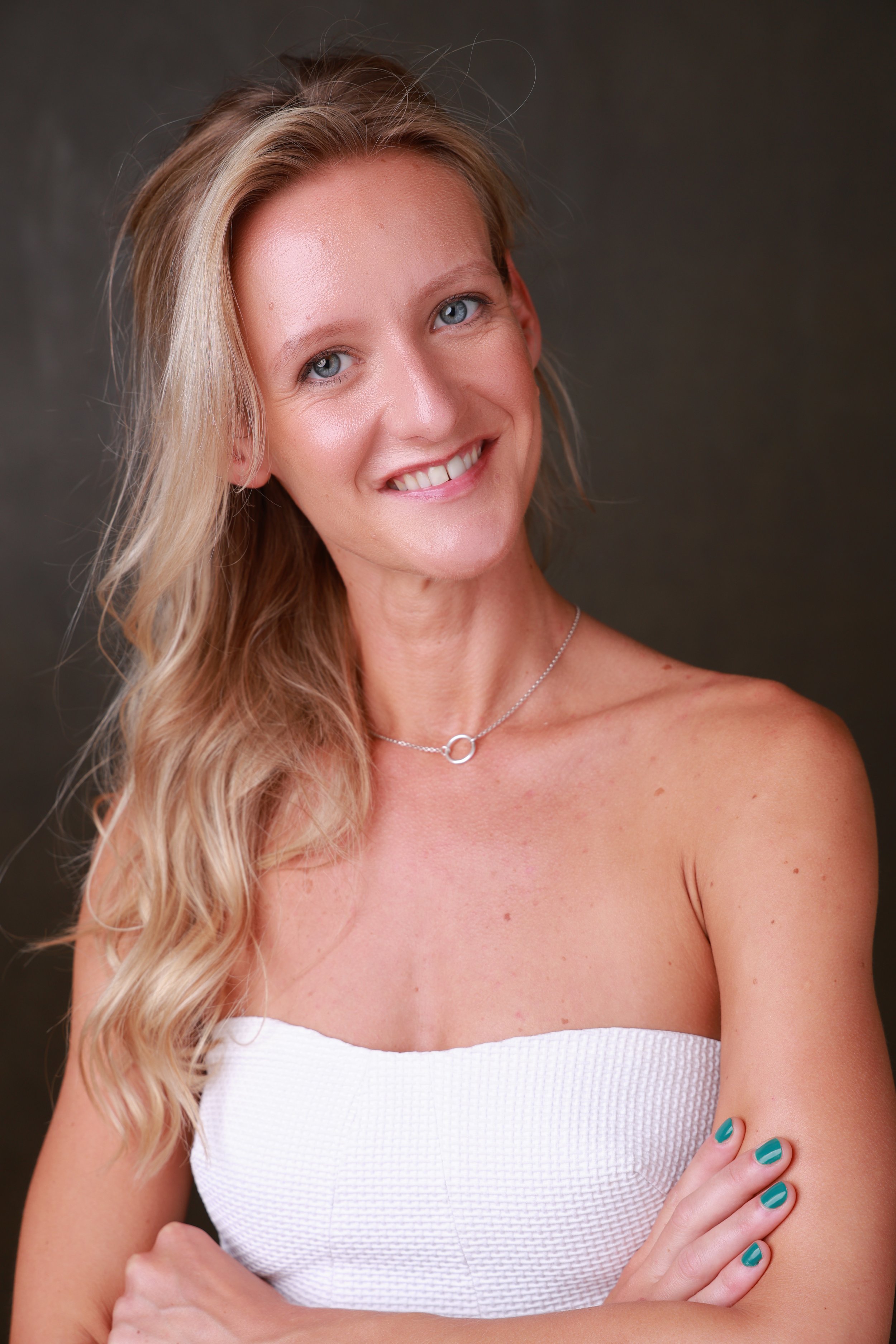 A smiling woman with long, wavy blonde hair wearing a white strapless top and a silver necklace, standing against a dark background.