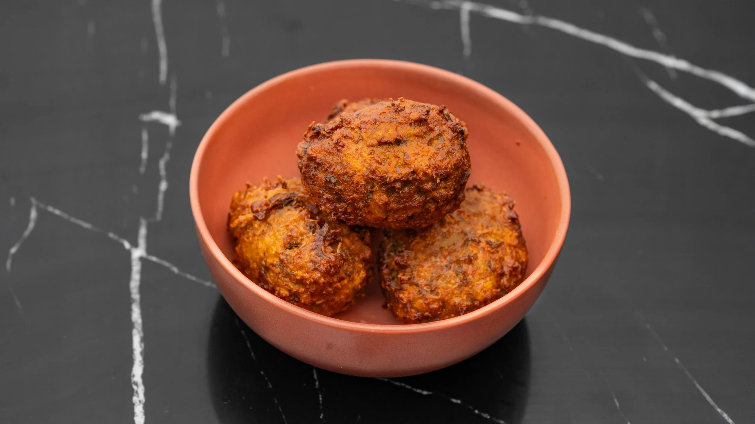 Trois boulettes de viande épicées dans un bol en terre cuite sur une surface noire avec des veines blanches.