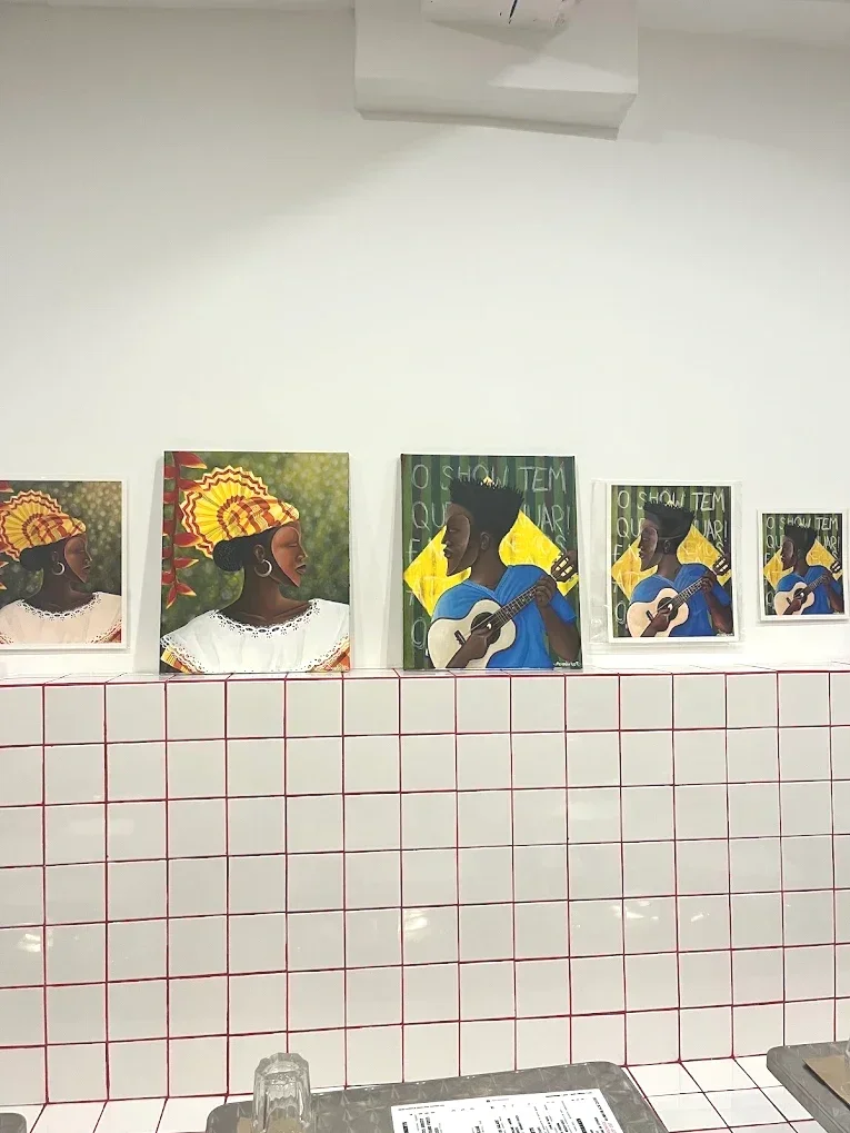 Quatre tableaux représentant deux femmes en profil portant un pagne coloré et un chapeau, et deux hommes jouant de la guitare, exposés sur un mur blanc avec une grille de pierre en dessous.