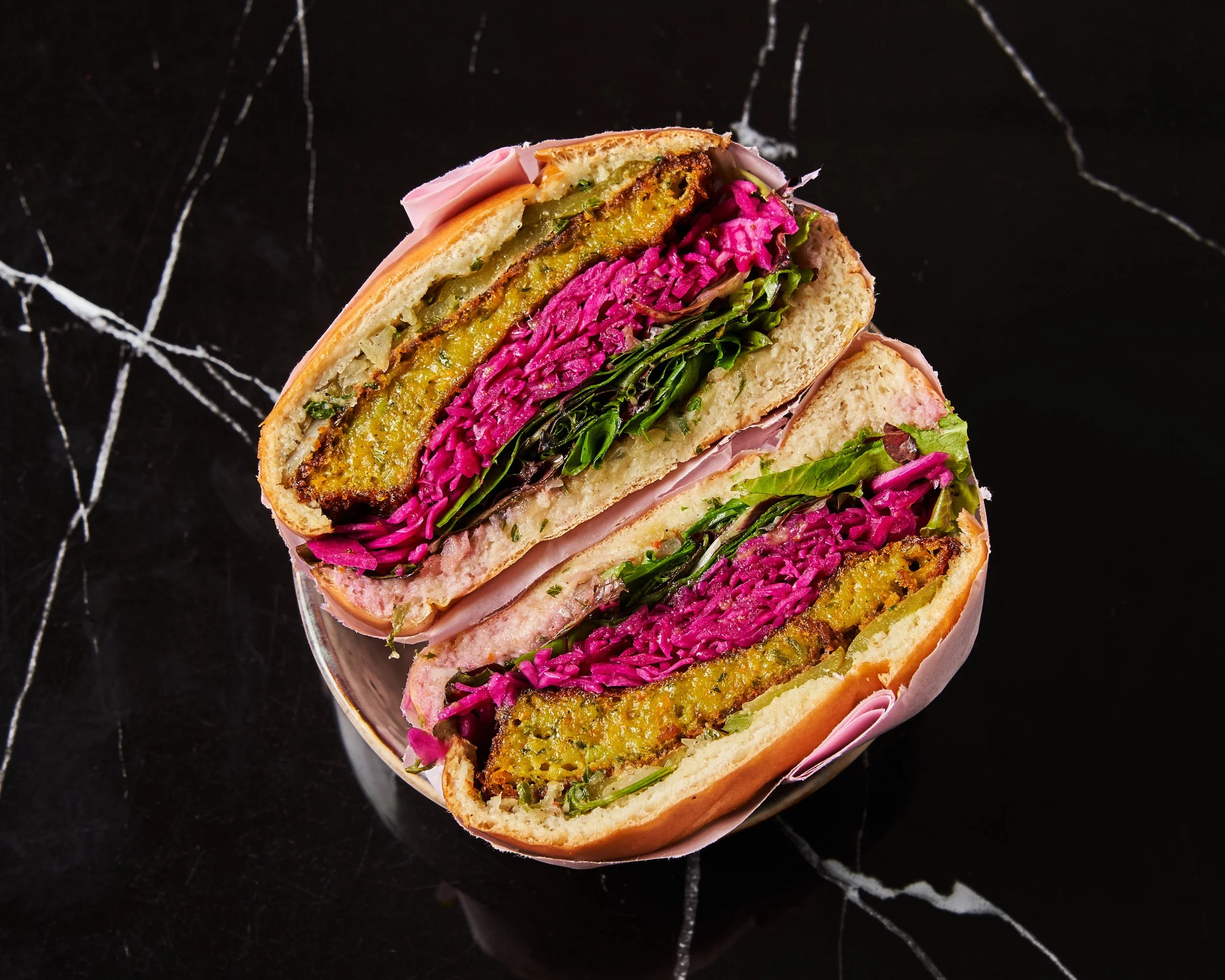 Sandwich coupé en deux montrant des couches de légumes colorés, de galette végétarienne, de jambon, et de salade, sur une assiette noire