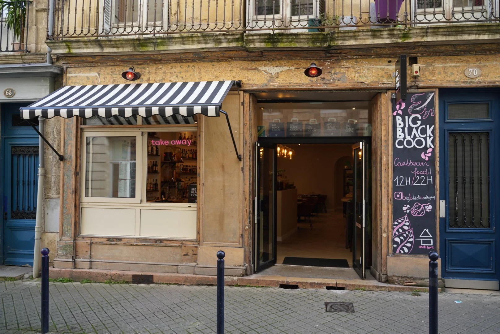 Une devanture de restaurant appelé 'Big Black Cook' avec une porte ouverte, un panneau noir avec des dessins de flamants roses et le nom du restaurant, ainsi qu'une fenêtre où l'on voit l'intérieur. Un panneau indique des horaires de 12h à 22h et mentionne la cuisine caraïbe.