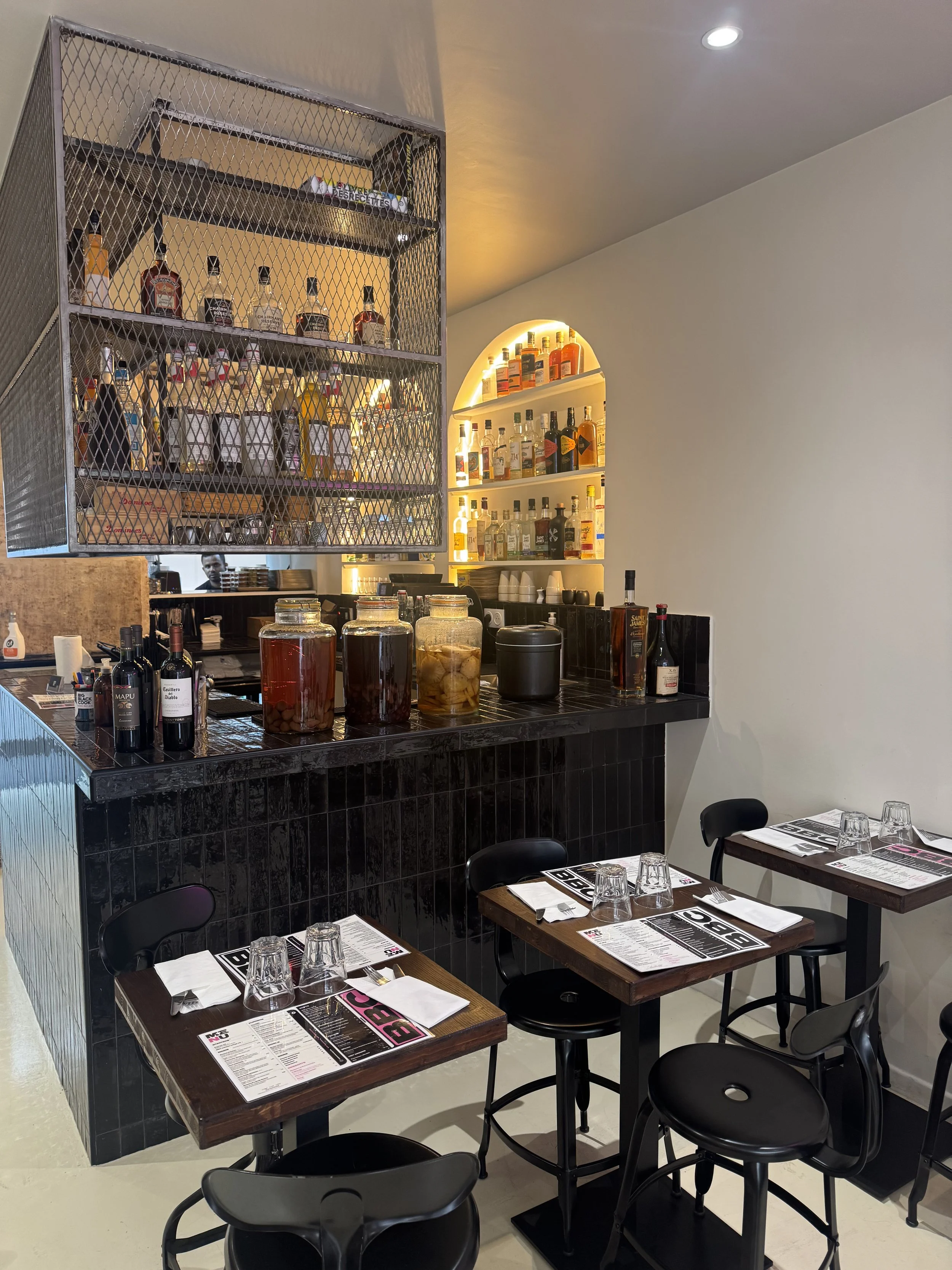 Une salle à manger de restaurant avec des tables en bois et des verres, en arrière-plan un bar avec des bouteilles d'alcool et des menus sur la table.