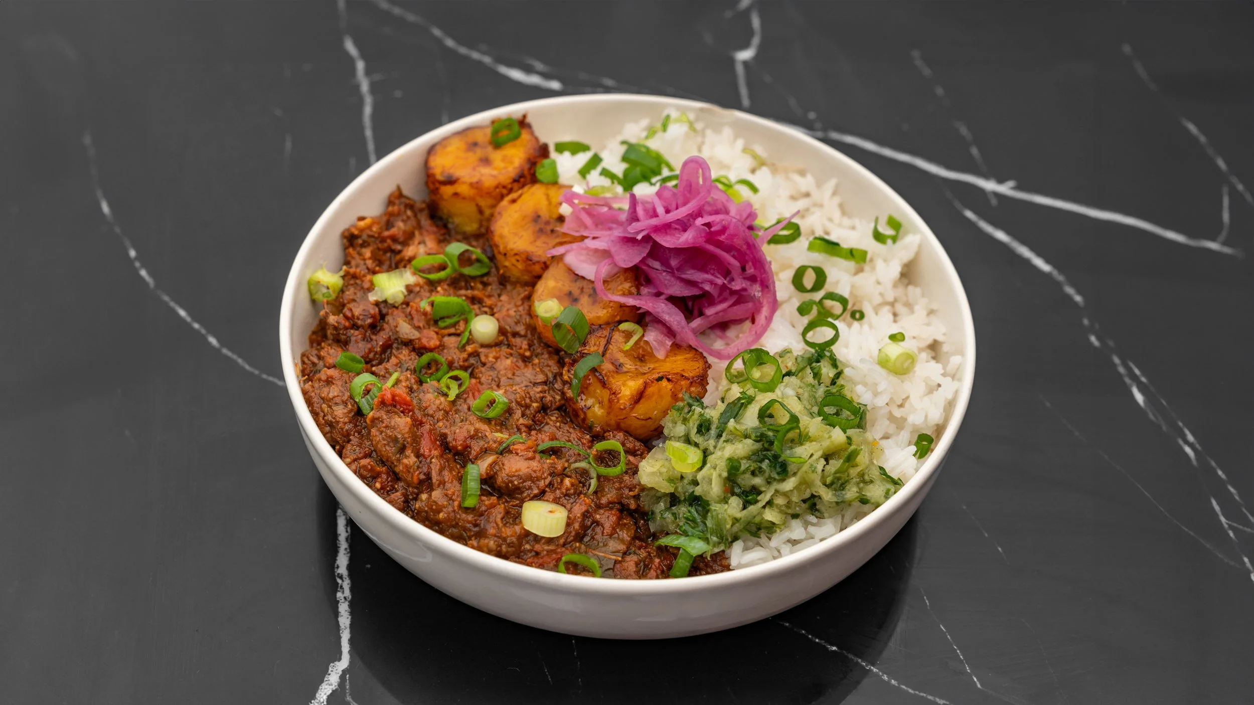 Bol de riz blanc avec un beef stew, des bananes plantains, de la salade d'avocat et de la choucroute, garnis d'oignons verts.