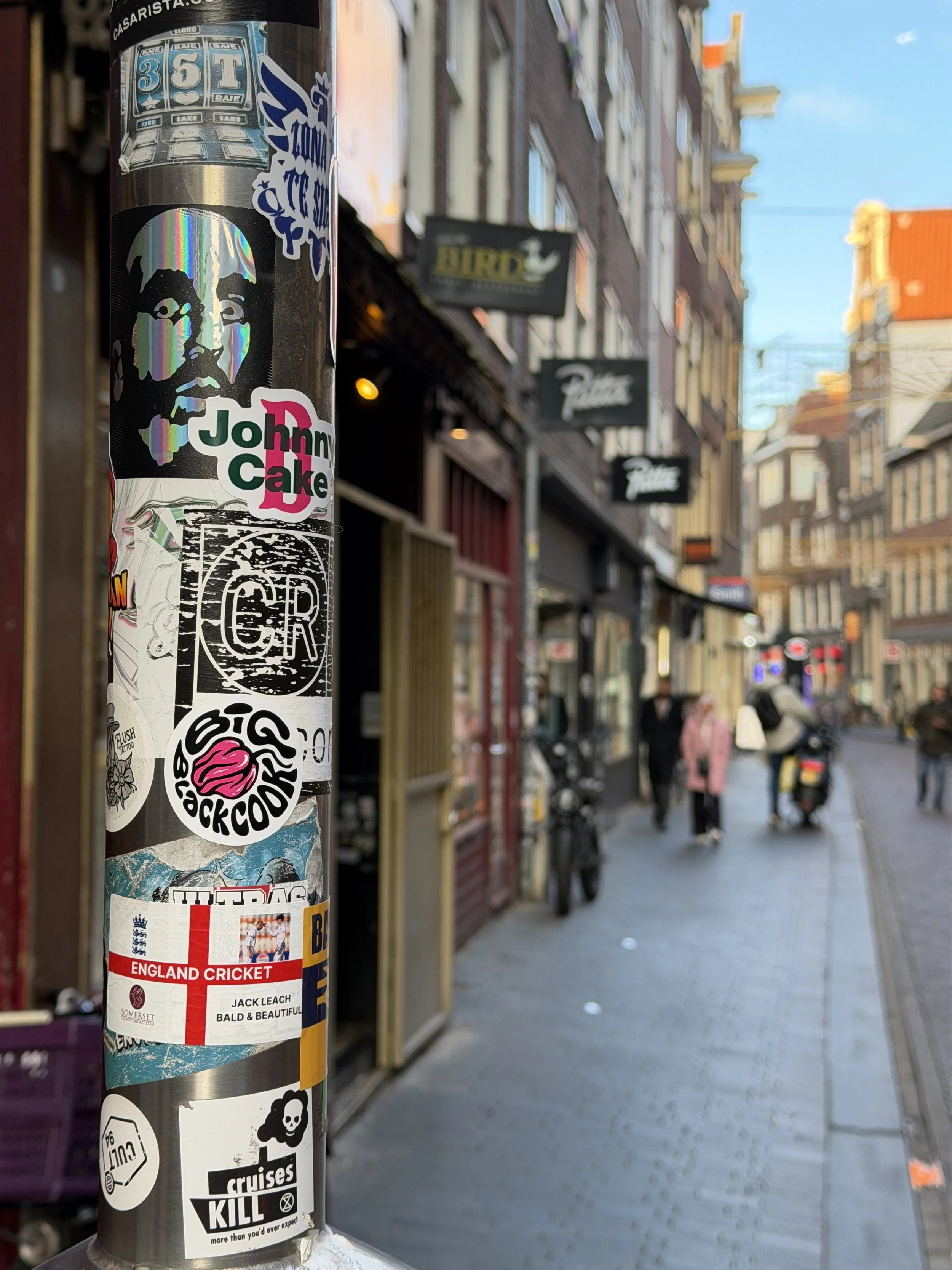 Sticker sur un poteau de rue avec diverses affiches et images, dont un portrait en style holographique et des textes, dans un quartier urbain animé