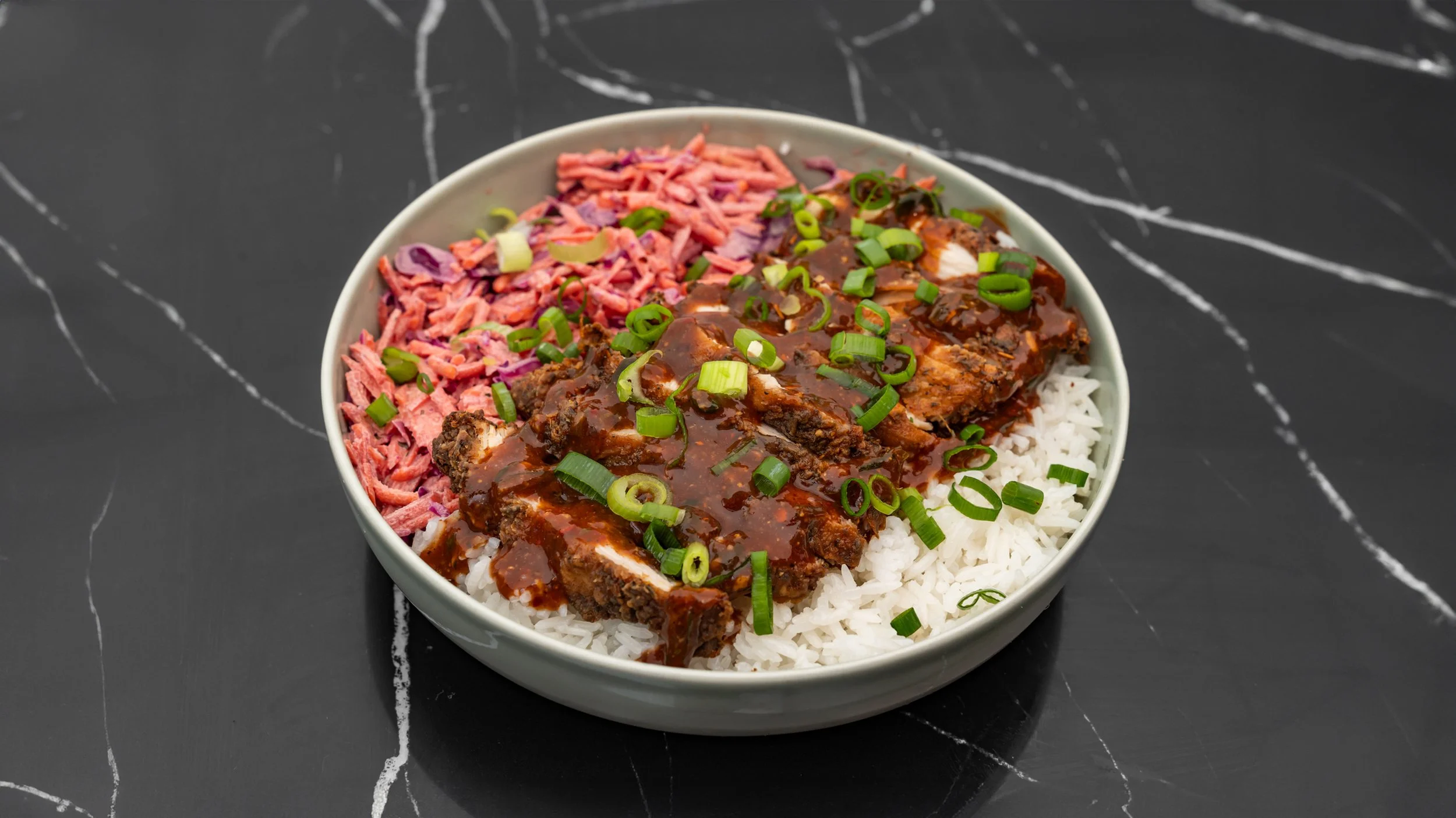 Bol de riz blanc garni de côtelettes de viande en sauce brune, de salade de chou, de coriandre et d'oignons verts, placé sur une surface sombre avec des veines blanches.