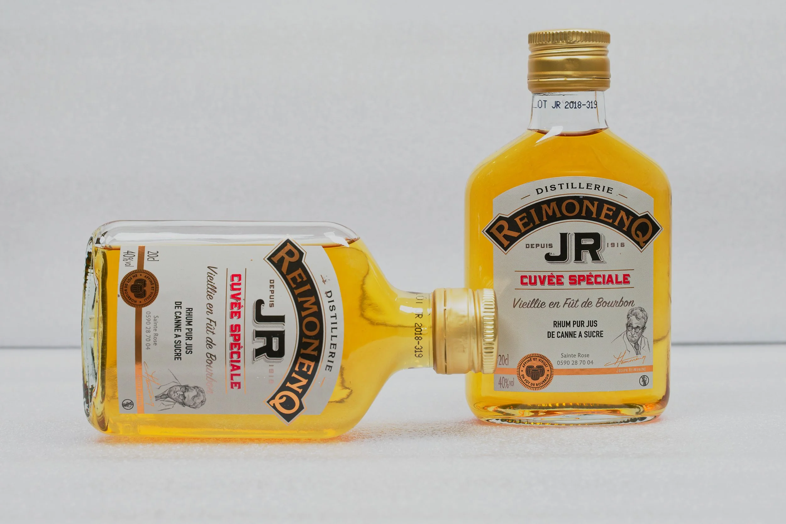 Deux bouteilles de rhum Reimonenq au premier plan, l'une est à l'envers, en dessous de l'autre, avec étiquettes indiquant qu'il s'agit d'un rhum vieux en fût de bourbon et d'une cuvée spéciale. Les bouteilles sont en verre transparent avec un liquide doré à l'intérieur, sur un fond neutre gris.
