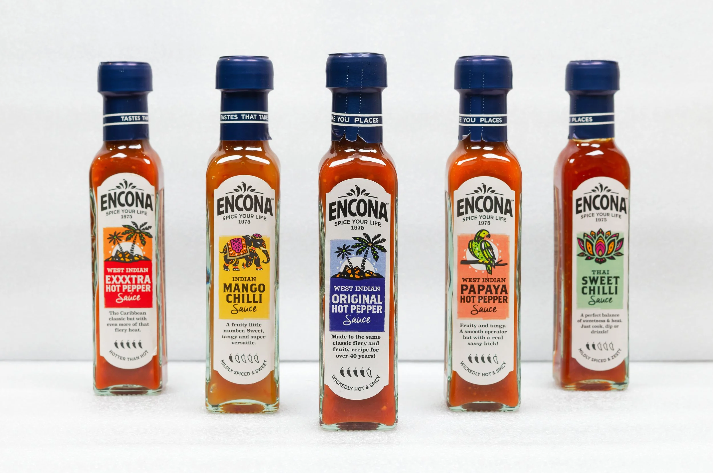 Cinq flacons de sauces piquantes Encona avec des étiquettes colorées représentant différentes saveurs, alignés côte à côte sur un fond blanc.