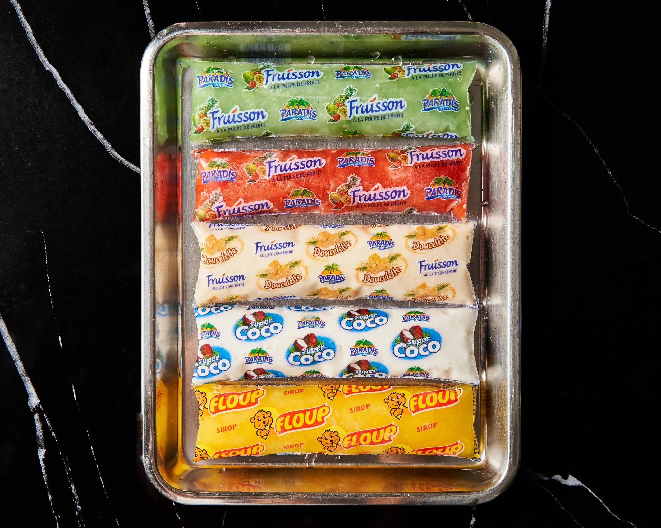 Différents empaquetages de yaourts aux fruits et au lait concentré, ordonnés dans un plateau métallique, comprenant des saveurs à base de fruits et de sirop.