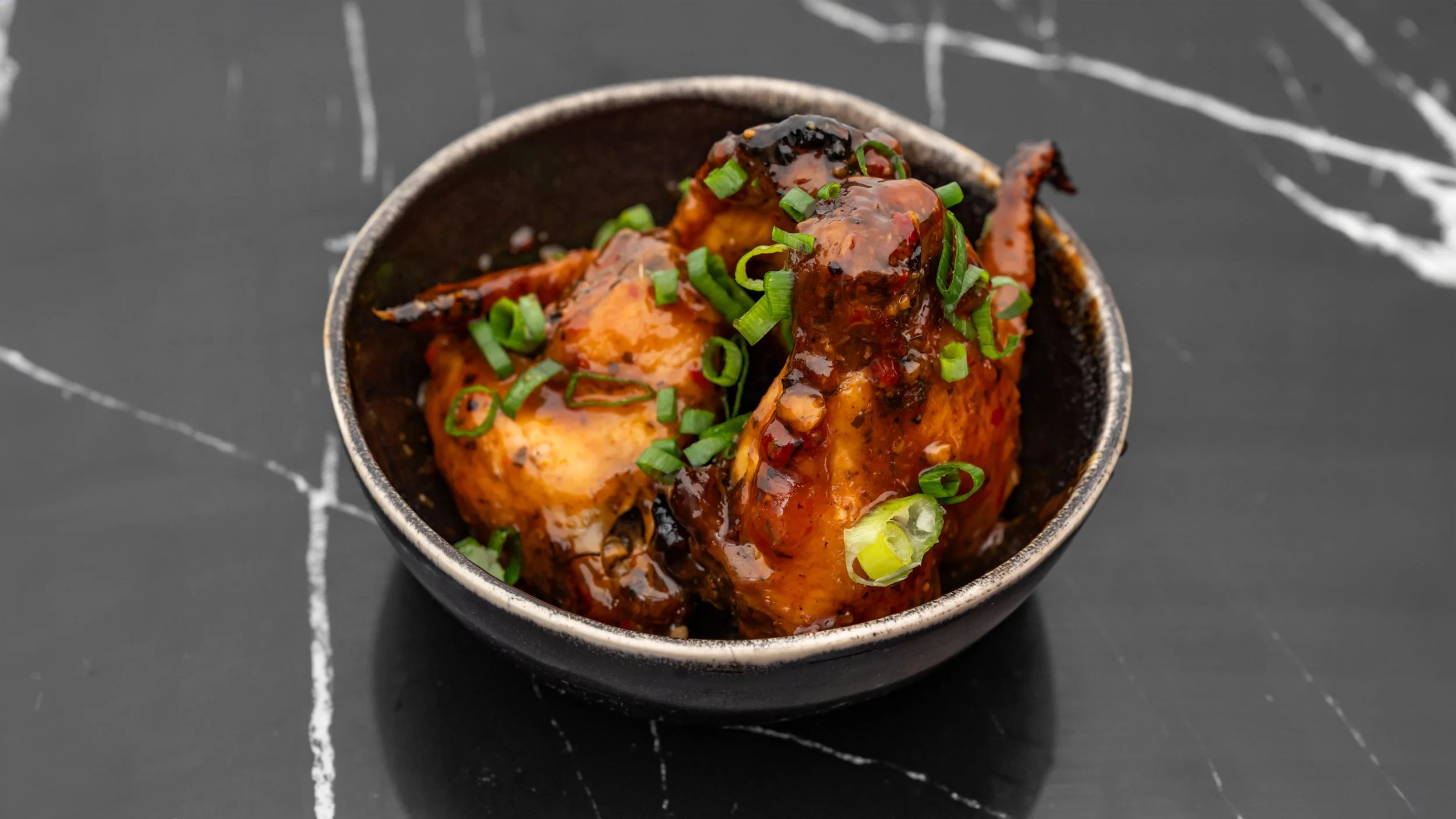 Poulet surmonté d'une sauce foncée, garni de ciboulette hachée, dans un bol noir, sur une surface noire avec des lignes blanches.