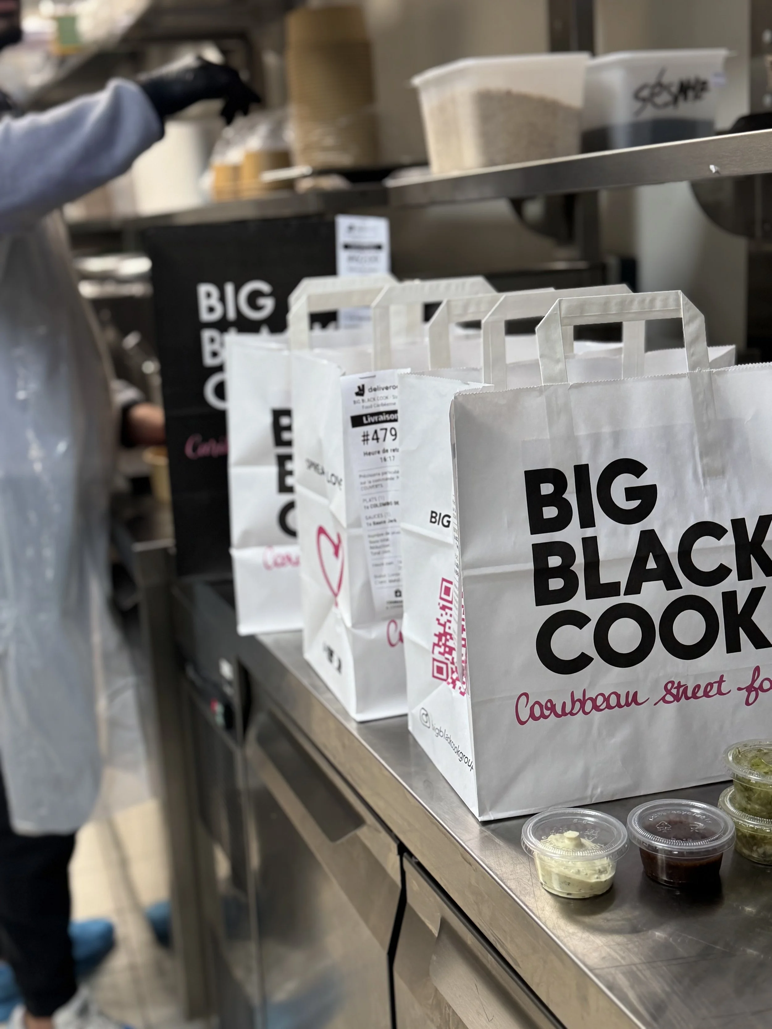 Sacs à emporter blanc avec écrit 'Big Black Cook' placés sur un comptoir de cuisine ou de restaurant, avec petits pots de sauces ou dips en-dessous.