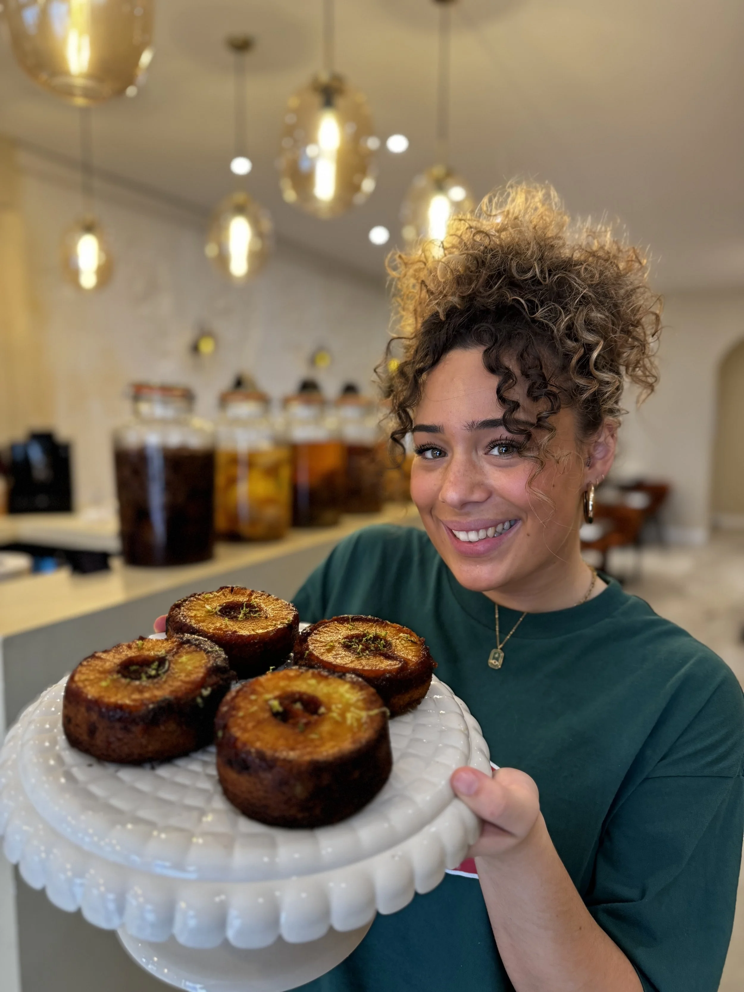 Jeune femme souriante avec cheveux bouclés tenant un gâteau aux fruits confits, devant un arrière-plan de distributeurs de boissons dans un café ou une boulangerie.