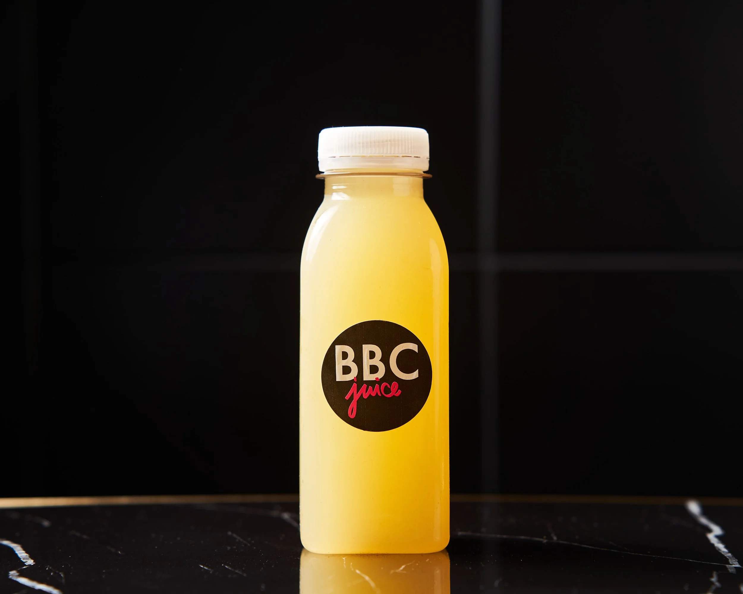 Bouteille de jus jaune avec un capuchon blanc, portant un logo noir avec le texte 'BBC juice', posée sur une surface noire.