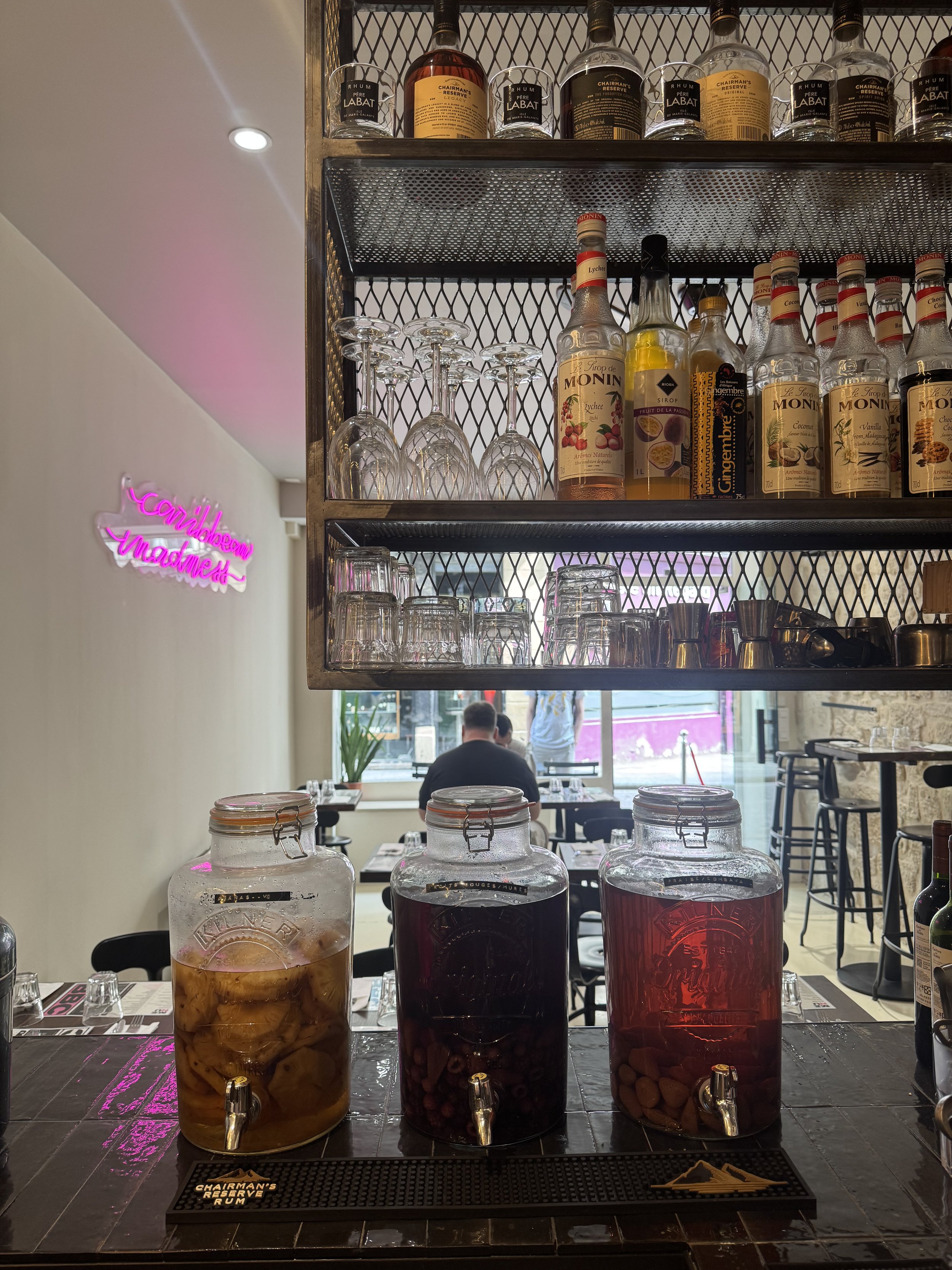 Des bouteilles de sirop et des jars contenant des fruits, avec des verres à vin suspendus au-dessus, dans un restaurant ou café.