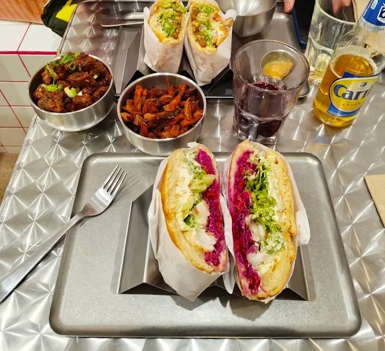 Sandwichs en sandwich à moitié ouverts avec du pain, de la laitue, du poulet et de la betterave, servis avec des accompagnements de viande, de frites, de boissons et de bière sur une table en métal.