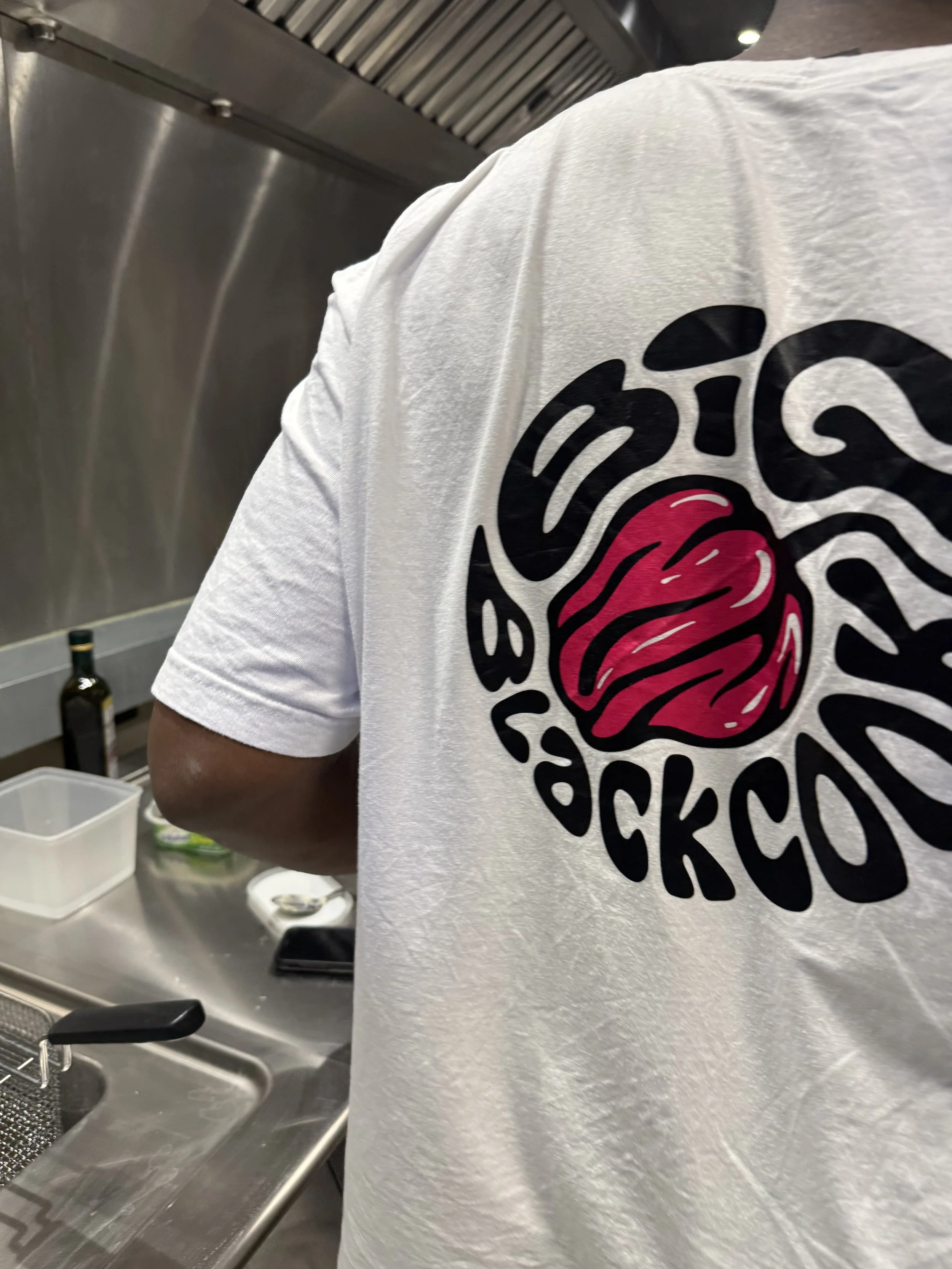 Un homme portant un t-shirt blanc avec un logo circulaire noir et rouge sur le côté. La scène se déroule dans une cuisine, avec une surface en acier inoxydable, une bouteille d'huile d'olive, un bol blanc et une boîte de rangement visibles en arrière-plan.