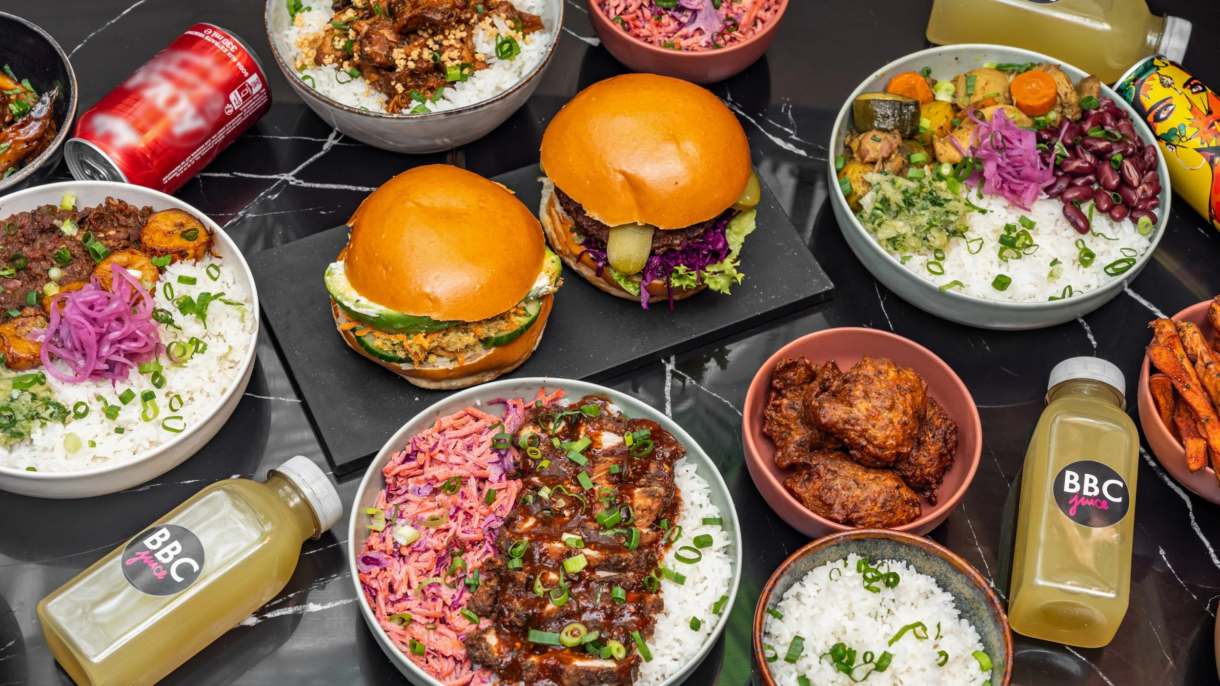 Variété de plats de cuisine africaine, include des bols de riz blanc, des sandwiches avec des pains briochés, des morceaux de viande en sauce, et des salades colorées, accompagnés de bouteilles de boissons