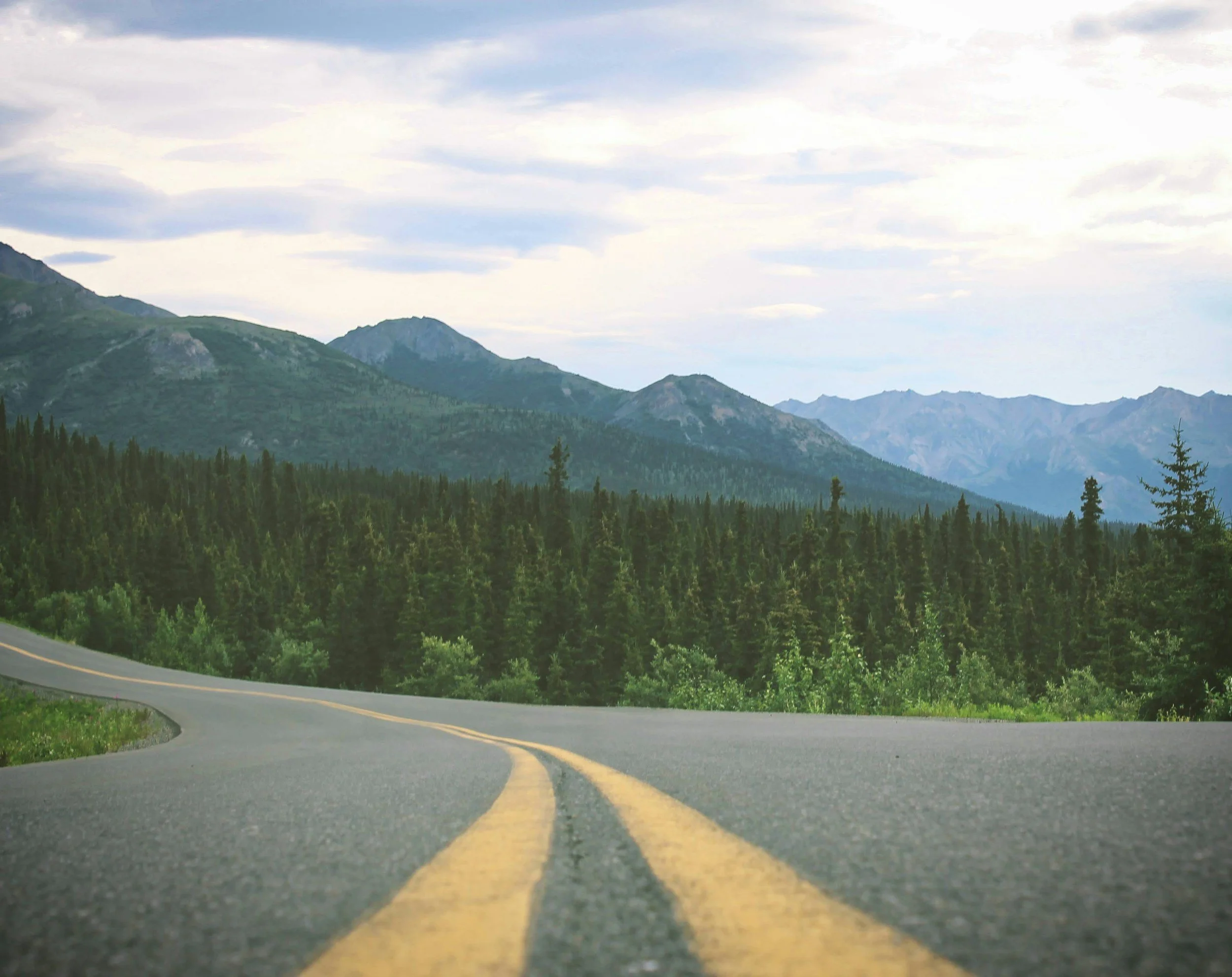 Curving mountain road with double yellow line, surrounded by green trees and mountains under cloudy sky.