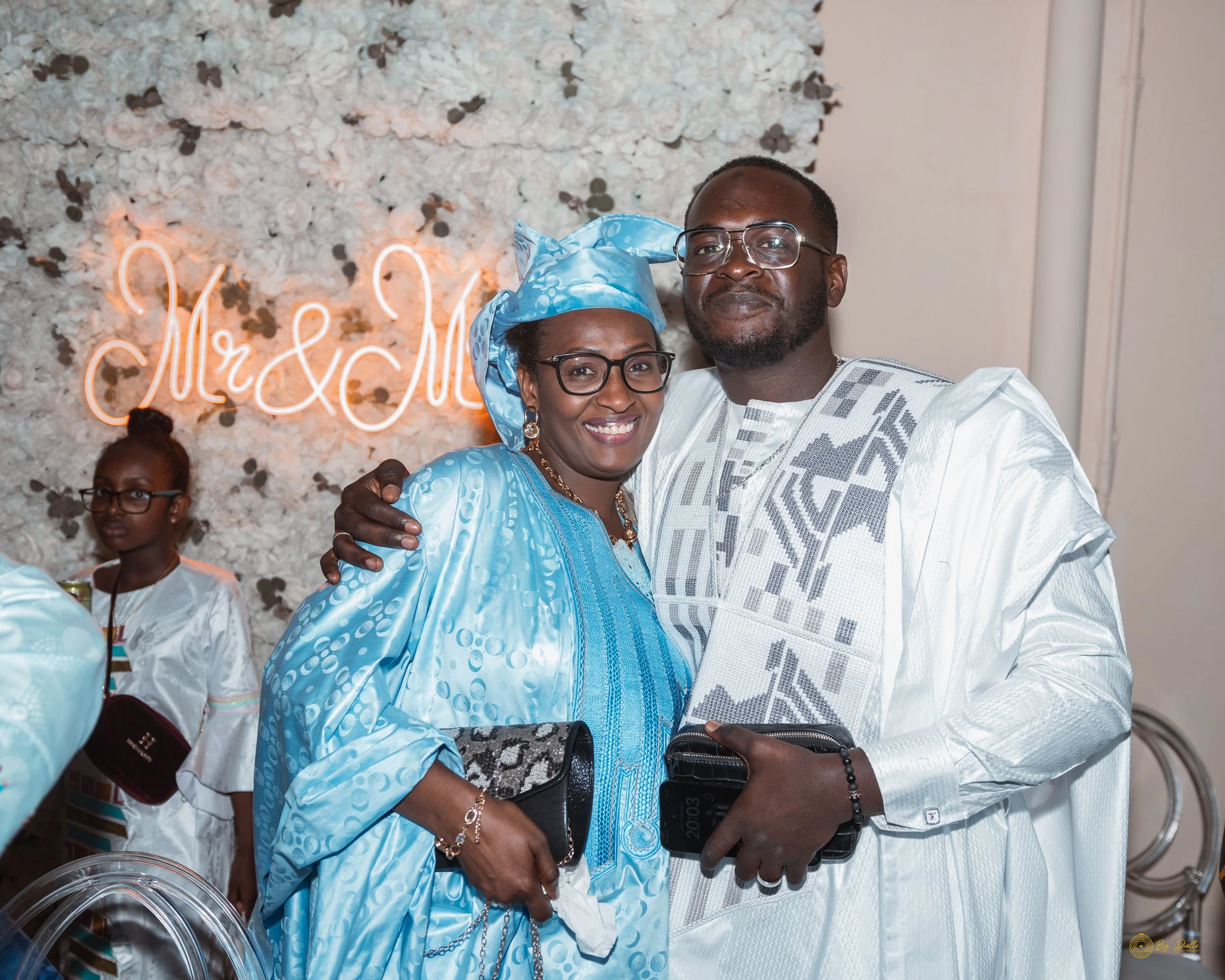 A happy couple dressed in traditional African attire at a celebration, with a floral backdrop and neon 'Mr & Mrs' sign, posing together and smiling.