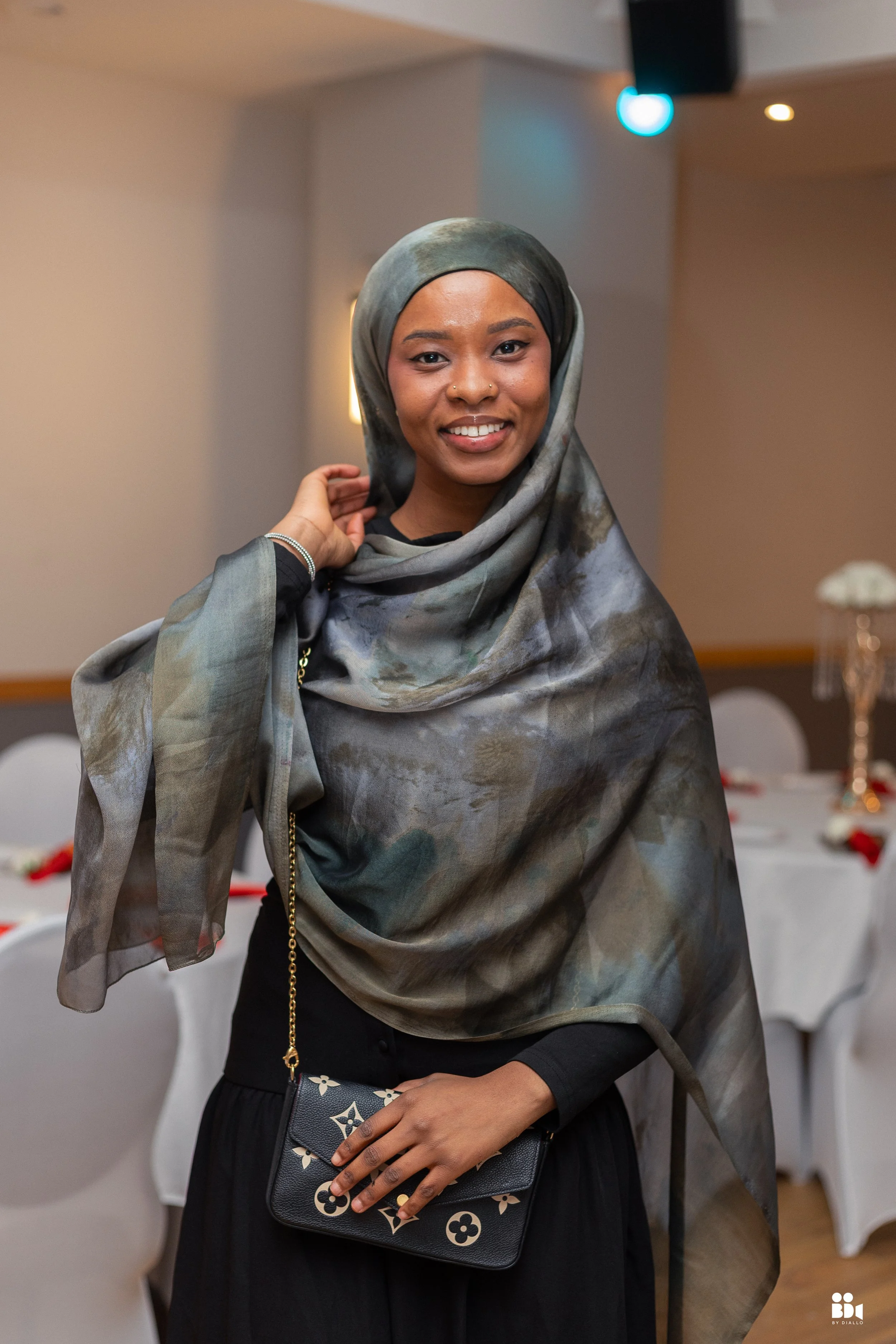A young woman smiling with a gray patterned hijab, holding a designer black clutch with floral patterns, in a decorated event space with tables and chairs in the background.