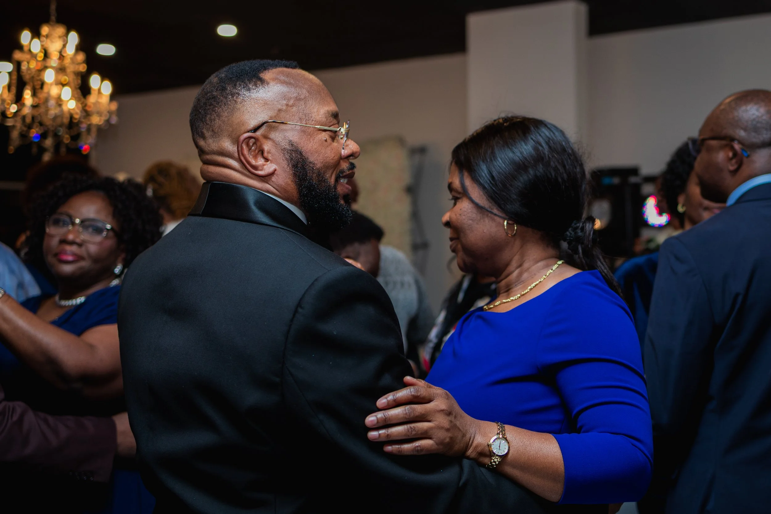 A man and woman are dancing closely at a formal event, smiling at each other, with other people dancing and socializing in the background.
