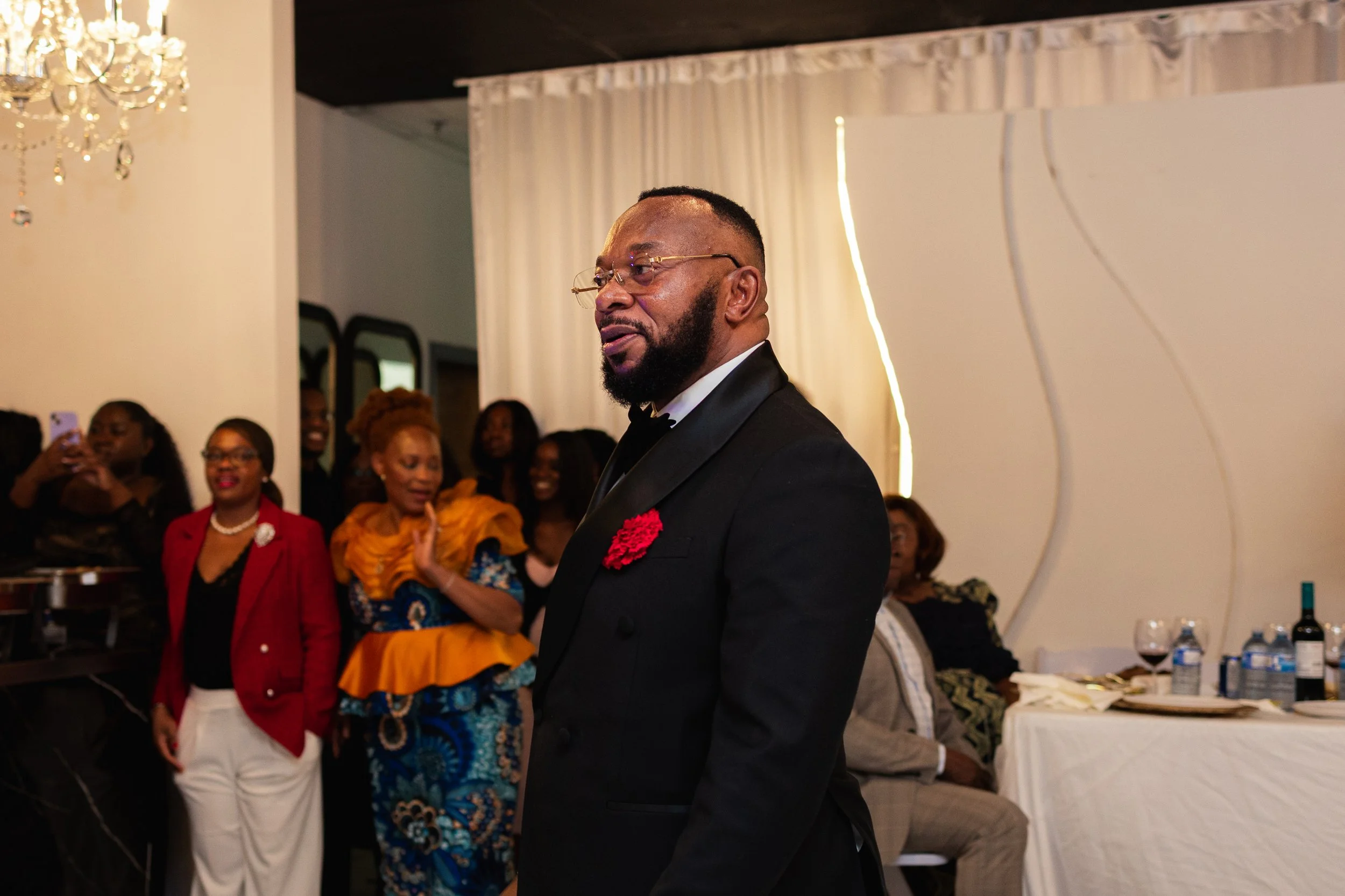 A man in a black tuxedo with a red flower on his lapel standing at a formal gathering, with women and a man seated and standing in the background, some smiling and taking photos, in an indoor event space with white curtains and a chandelier.