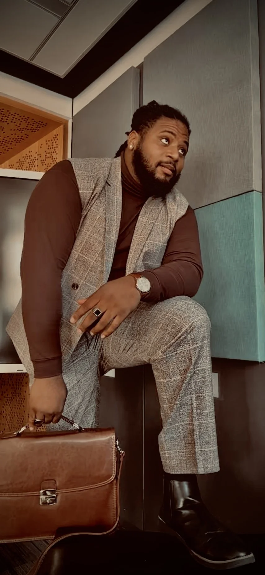 Jonathan Gilbert with a beard and dreadlocks is dressed in a brown turtleneck, plaid gray pants, and a gray vest. He is sitting in a modern interior, holding a brown leather briefcase, looking thoughtfully to the side.