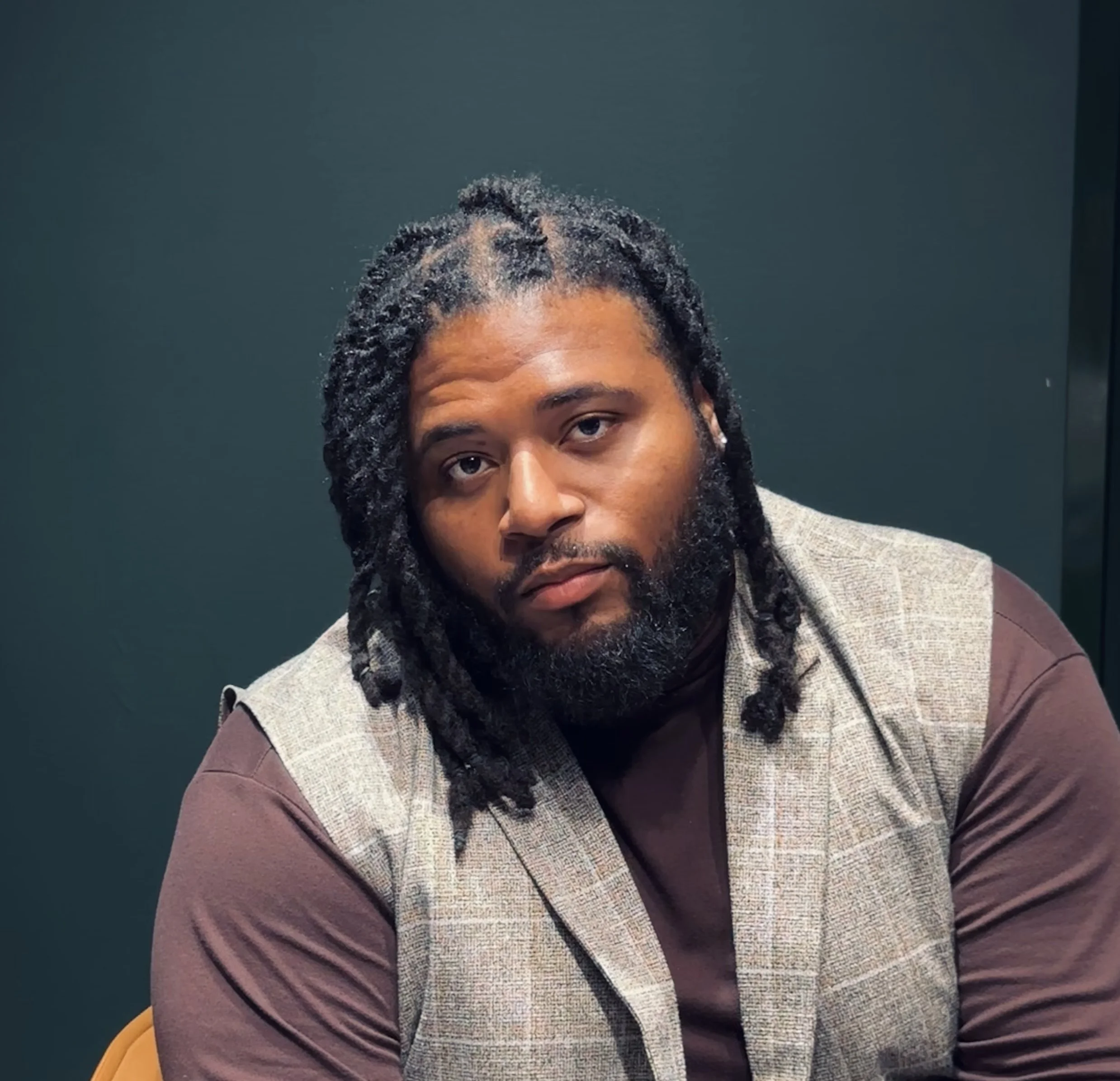 Jonathan Gilbert with dreadlocks and a beard sitting against a dark green background, wearing a plaid vest over a brown shirt, looking at the camera.