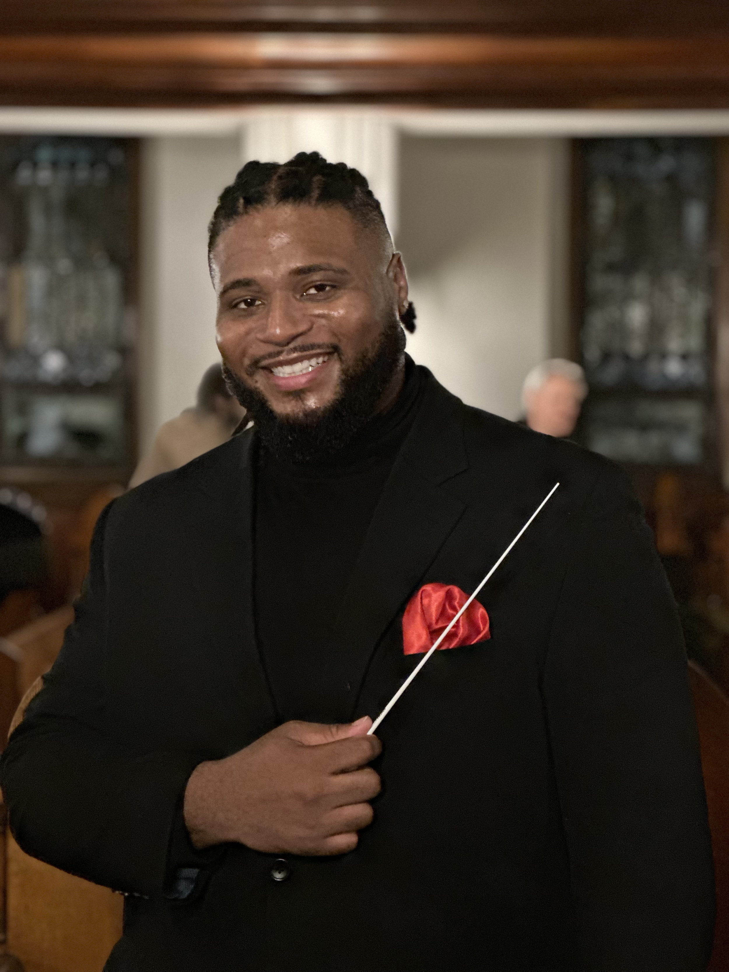 Jonathan Gilbert has a beard and locs, dressed in a black suit with a red pocket square, holding a conductor's baton in a warmly lit room with blurred background of other people.
