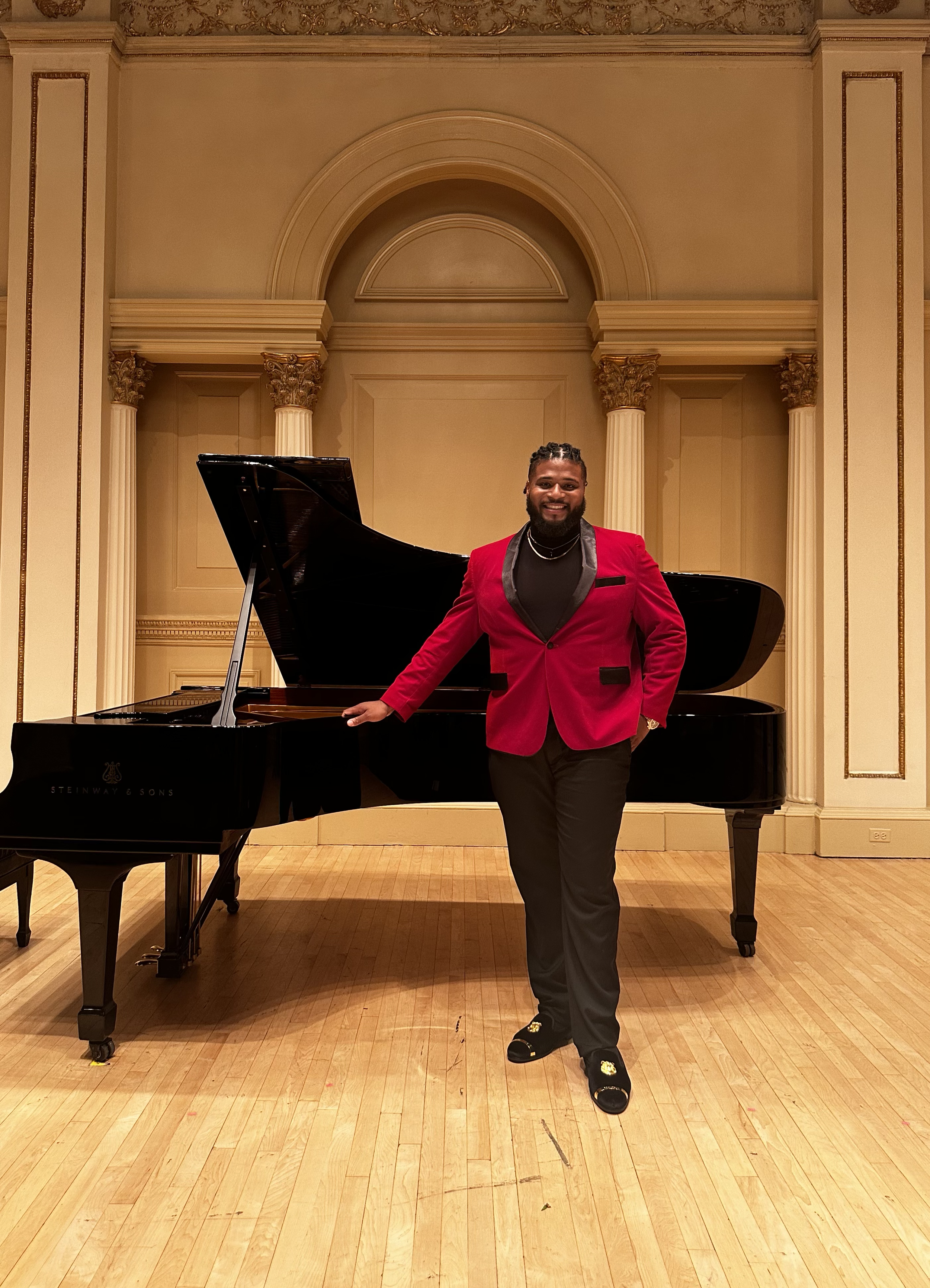 Jonathan Gilbert in a red blazer and black pants standing beside a black grand piano in Carnegie hall.
