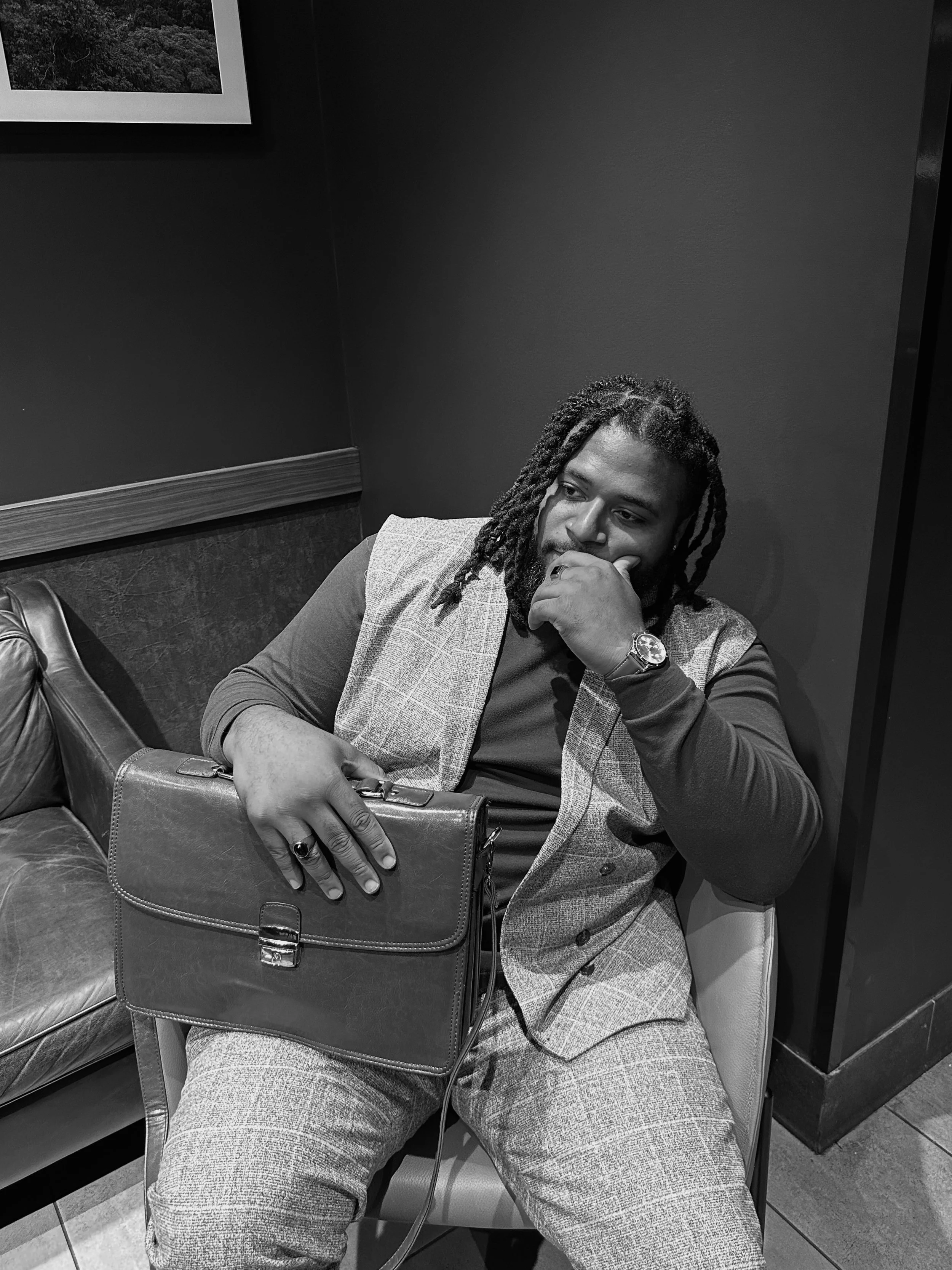 Jonathan Gilbert with dreadlocks sitting on a chair in a black and white photo, holding a briefcase, with a hand on his face, wearing a checkered vest and watch.